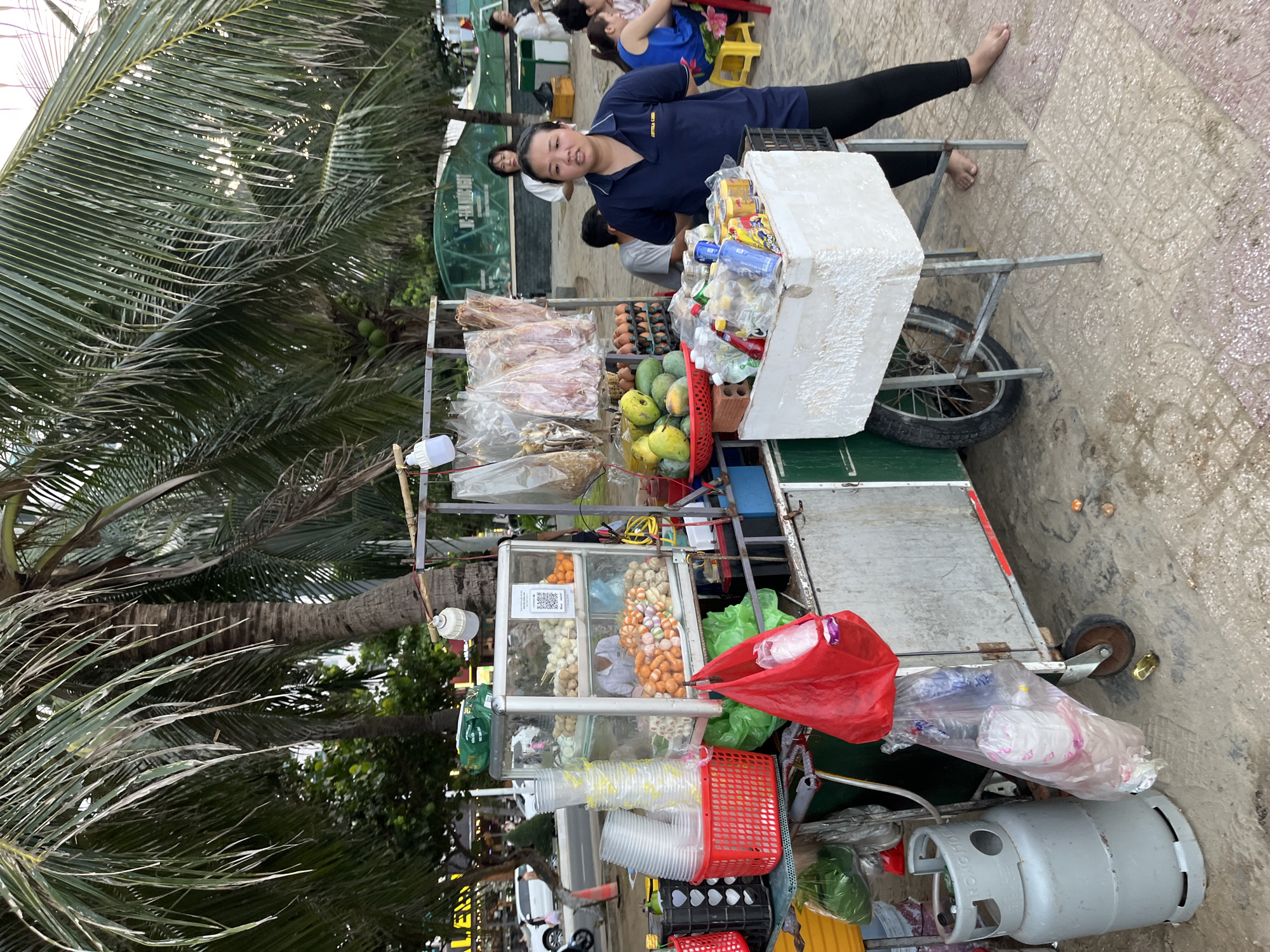 A street food cart is displayed in Bac Nha Trang Ward, Vietnam. The cart is laden with various food items including eggs, mangoes, dried fish, and assorted snacks. A woman stands behind the cart, wearing a navy blue polo shirt and black leggings. Two other women and several men are visible in the background. Palm trees are present, suggesting a coastal or tropical environment. The ground is paved with tiles, and there's a light sandy texture. A gas cylinder is also visible next to the cart.
