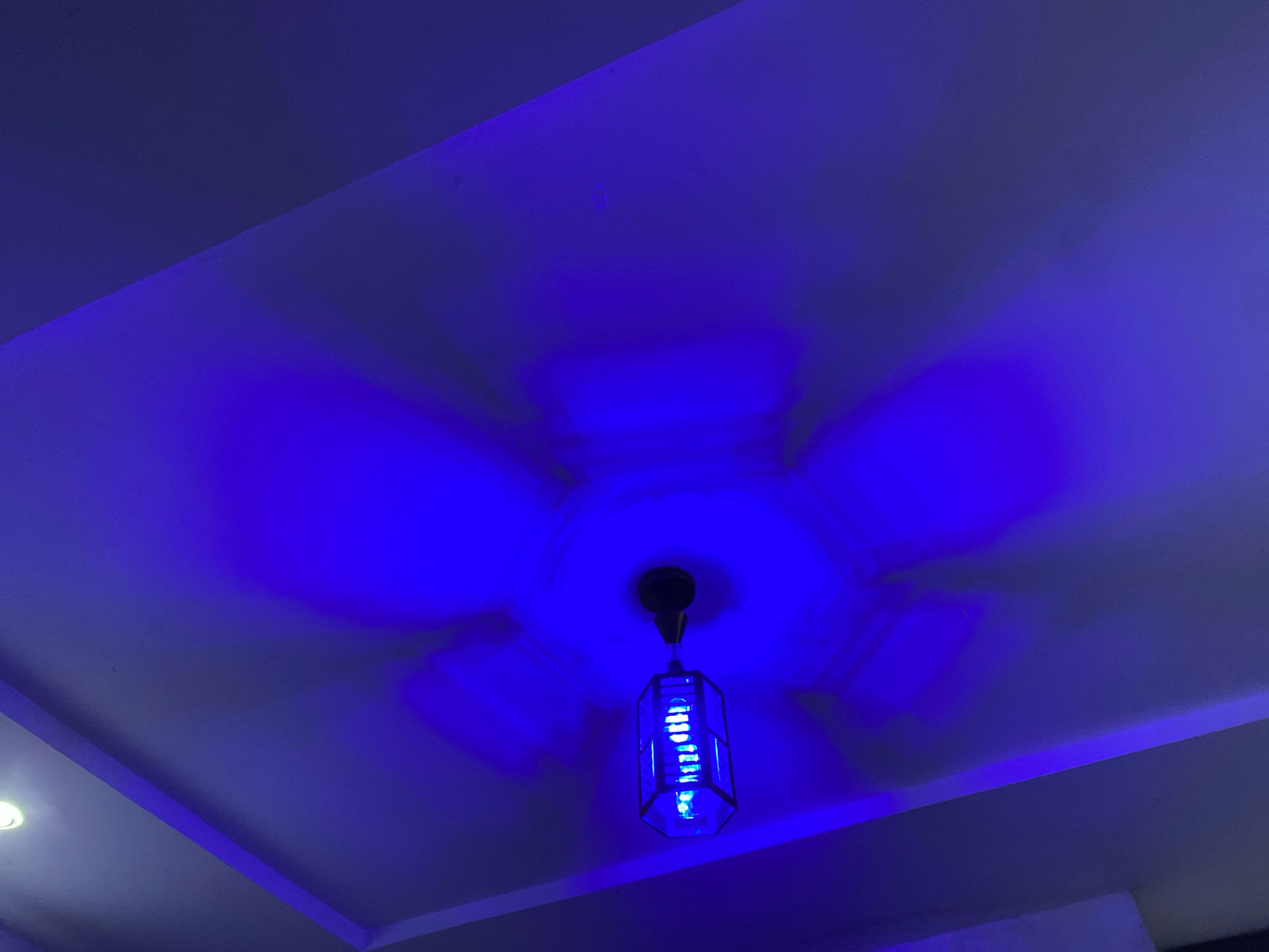 The image shows a blue illuminated ceiling in Shangisha, Nigeria. A modern hanging lamp with a hexagonal glass shade, emitting a bright blue light, is suspended from the center of the ceiling. The ceiling has a decorative recessed design, and the blue light casts a diffused glow, creating shadow patterns that extend outwards from the lamp, suggesting the presence of ceiling fan blades above it, though they are not directly visible. The overall impression is of a contemporary interior space illuminated by atmospheric blue lighting.