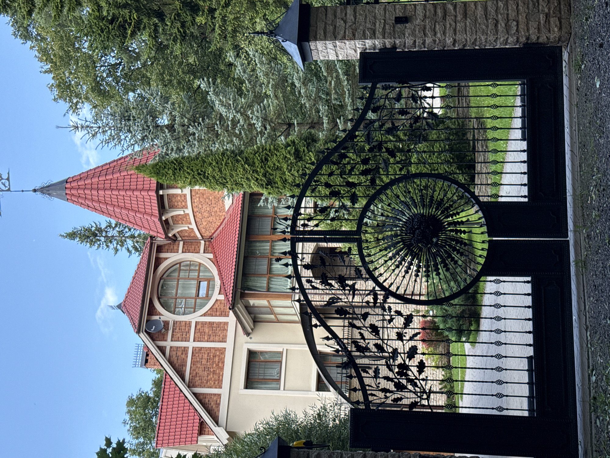 The image shows the ornate wrought-iron gates of a large, multi-story house in Krasnoye Selo, Russia. The house features a red tile roof, brick accents, and a prominent circular window. The gates are adorned with a decorative motif of leaves and branches, and a large sunburst-like design in the center of one gate. The driveway leading to the house is paved with gray stones. Tall evergreen trees and other foliage surround the property, suggesting a suburban or rural setting. The sky is blue with a few wispy clouds. The scene is captured from a low angle, emphasizing the grandeur of the gates and the house. There are no people visible in the image.