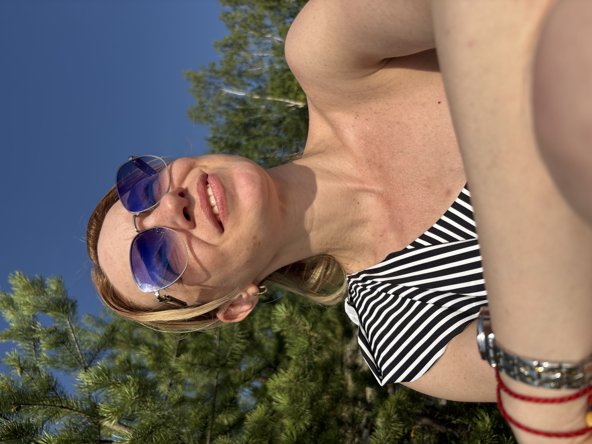 A woman with blonde hair pulled back in a ponytail, wearing blue-tinted sunglasses and a black and white striped top, smiles as she looks up at the sky. She has on a hoop earring in her left ear and a watch on her left wrist, along with a red string bracelet. The background consists of green pine trees, other foliage, and a clear blue sky, suggesting an outdoor setting, possibly in Shuiskaya Chupa, Russia.