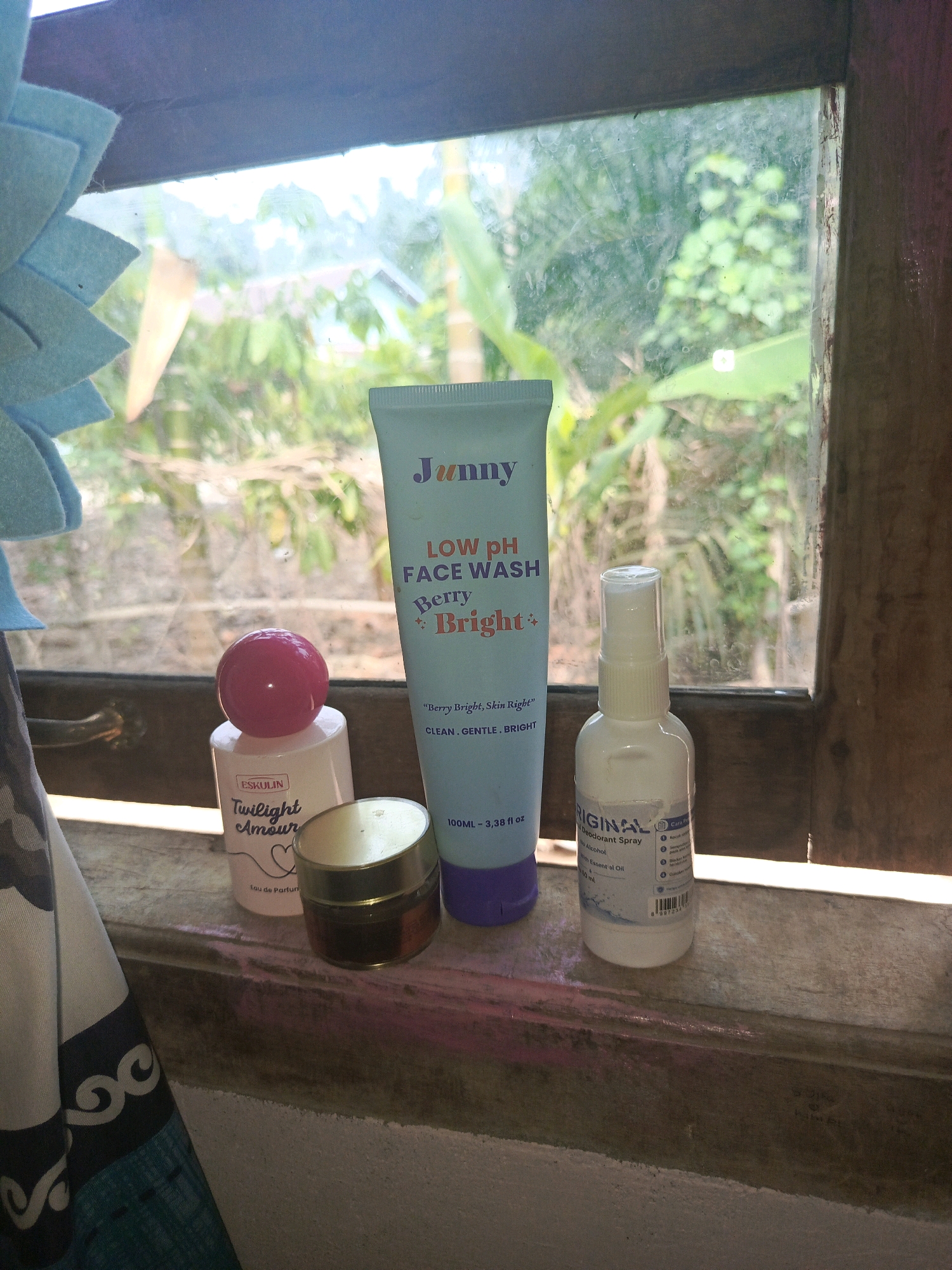 Four cosmetic products are arranged on a wooden ledge in front of a window. From left to right, the first item is a small, white bottle with a pink spherical cap, labeled "ESKULIN Twilight Amour Eau de Parfum". Next to it is a small, gold-colored jar with a clear bottom. To the right of the jar is a large, light blue tube of "Junny LOW pH FACE WASH Berry Bright". Finally, on the far right, is a small, white spray bottle labeled "ORIGINAL Deodorant Spray". The window behind the items shows a blurred view of lush green foliage and a hint of a building, suggesting an outdoor environment. The wooden ledge appears weathered. The scene is lit by natural light coming through the window. The overall impression is that of a personal collection of toiletries displayed in a domestic setting, likely in the city of Megang Sakti I, Indonesia, given the context.