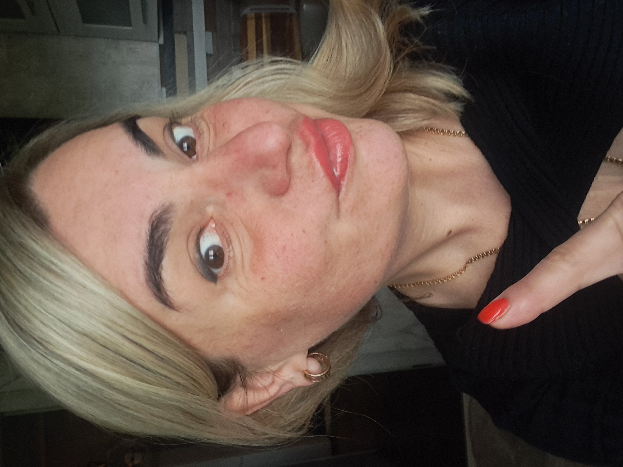 A woman with blonde hair, wearing a black top and a gold necklace, is posing for a selfie. She has red lipstick and red nail polish. Her ear has a gold hoop earring. The location is indoors. Elektrostal, Russia is not identifiable from the image.