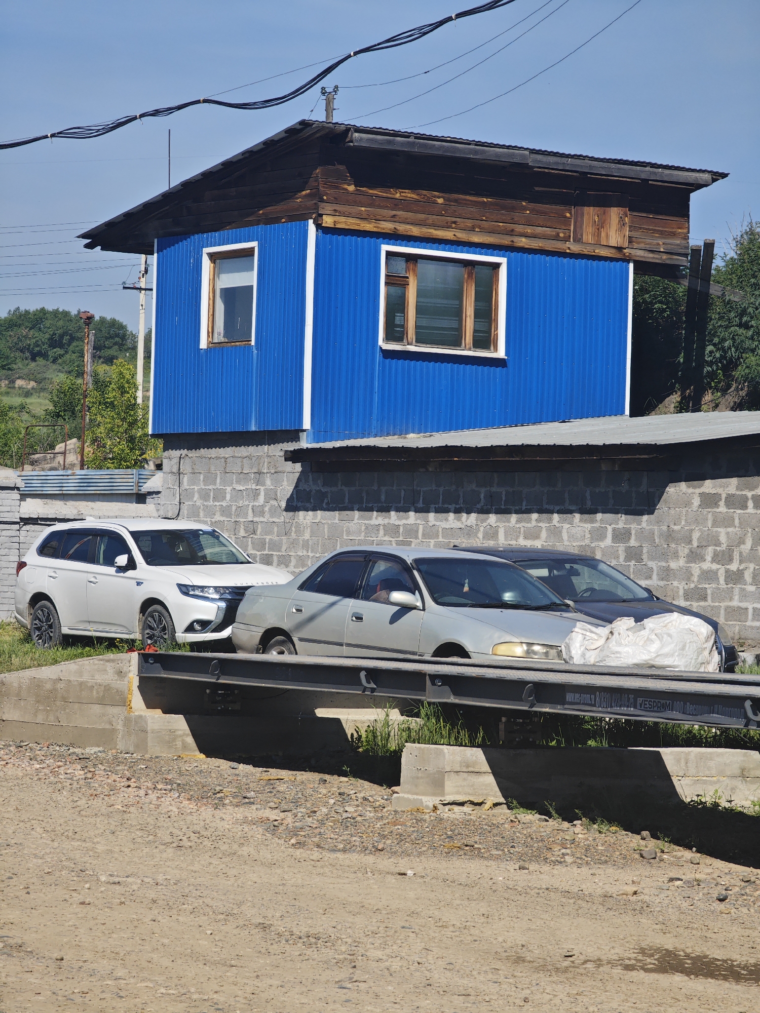The image depicts a bright, sunny day in Krasnoyarsk, Russia. The scene features a weathered, two-story structure with a blue corrugated metal lower section and a wooden upper section. Two windows with wooden frames are visible on the blue portion. A dark, sloped roof tops the building. Power lines crisscross the clear blue sky above.
In front of the structure, a gray cinder block wall runs horizontally across the frame. Several cars are parked in front of this wall. On the left, a white SUV is parked on a grassy area. To its right, a silver sedan is partially visible, followed by a dark-colored car covered with a large white tarp.
Beneath the cars and wall, a concrete bridge or overpass is visible, with a metal railing along its edge. The area in the foreground is a dusty, gravelly road. The overall impression is of a somewhat rustic and utilitarian setting, likely on the outskirts of a town or in a rural area, given the visible construction materials and natural elements like trees and grassy slopes in the background. The time of day appears to be midday, given the strong sunlight and shadows.