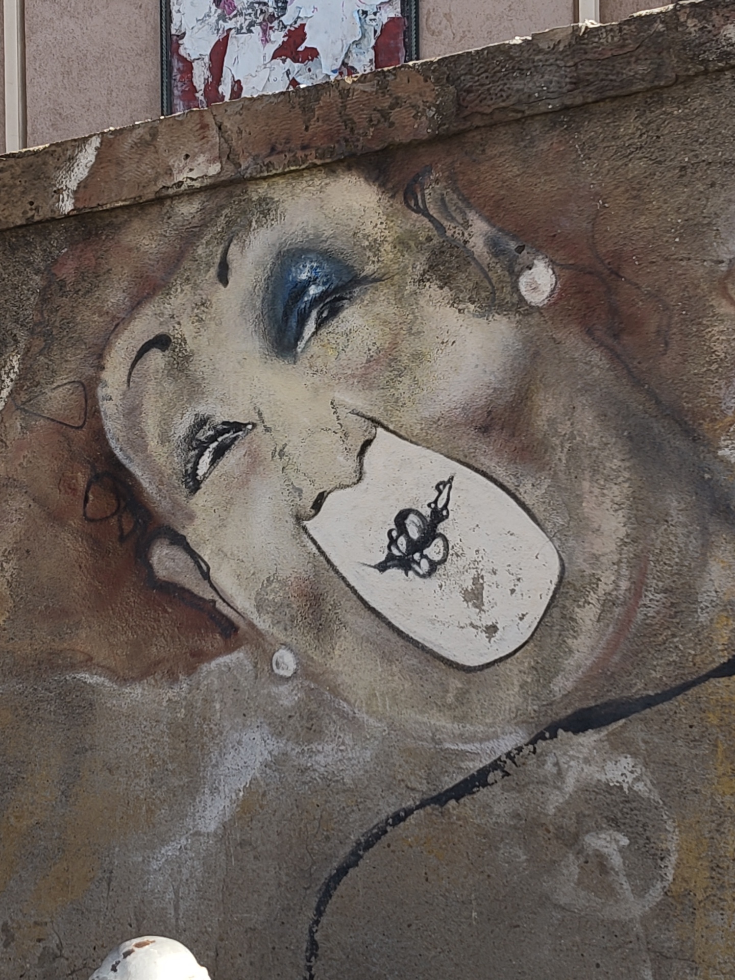 The image shows a weathered mural on an outdoor wall in Cagliari, Italy. The mural depicts a stylized face with heavy makeup, one eye closed in a wink. The mouth is open with a white rectangle inside bearing a dark, abstract design resembling a bug or stylized lips. The mural appears aged, with cracks and signs of wear, and is executed in shades of brown, tan, black, and white. The wall is made of rough stone. Above the mural, a section of the wall and a framed piece of what appears to be torn posters are visible. A small white round object is visible in the lower left.