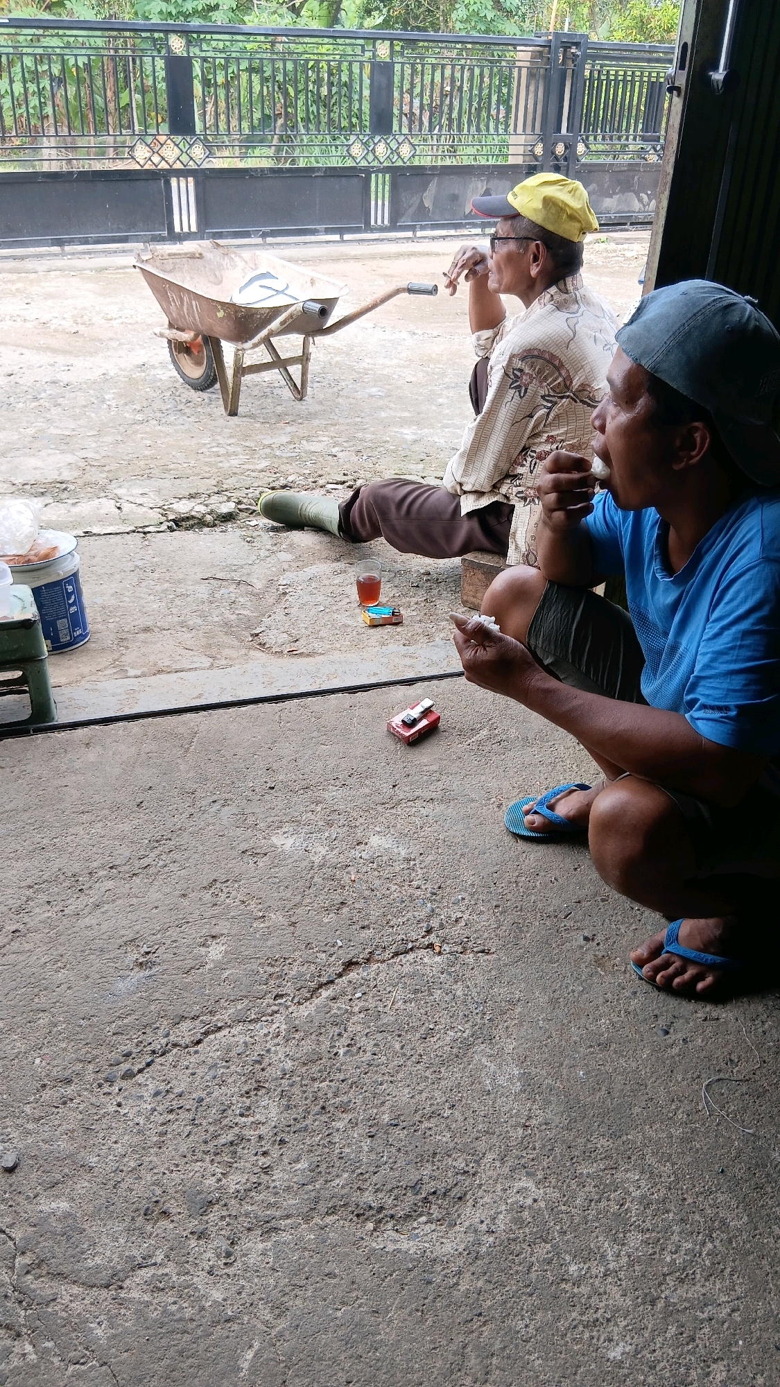 Two men are seated on a concrete surface outside a building in Sungai Tarab, Indonesia. The man on the left wears a yellow cap, glasses, and a patterned shirt. He is holding a cigarette to his mouth and appears to be smoking. He is also wearing brown pants and green boots. A wheelbarrow is positioned to his left. The man on the right wears a blue baseball cap and a blue t-shirt. He is holding a small white object to his mouth, possibly food or a piece of tissue. He is wearing shorts and blue flip-flops. A pack of cigarettes and a small glass of dark liquid are on the ground between the two men. A small red and silver object, possibly a toy or a lighter, is also on the ground near the man on the right. The background shows a black metal fence and lush green foliage. The scene appears to be a casual outdoor setting.