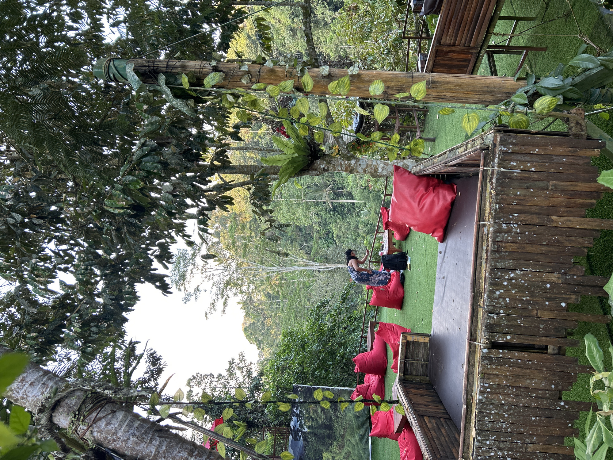 A person stands in a natural outdoor seating area in Tembuku, Indonesia, surrounded by lush greenery and trees. The seating area features bright red beanbag chairs scattered across a vibrant green carpeted floor. In the foreground, a rustic, open-air structure made of bamboo and wood is visible, with a dark interior and a wooden railing. To the right, a wooden platform with seating is also present. The person, wearing a dark patterned dress, appears to be interacting with a table and a black backpack. The background reveals a dense forest with a bright sky peeking through the canopy. The overall ambiance suggests a tranquil and natural environment, likely a cafe or recreational spot.
