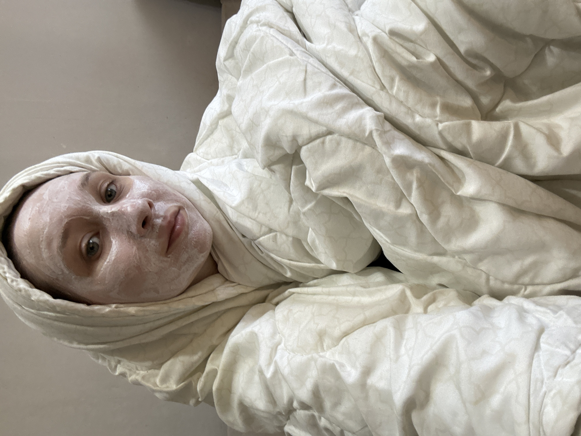 The image shows an individual indoors, likely in Chaikovsky, Russia, engaged in a self-care routine. The primary subject is a person, possibly female, with a white face mask applied evenly across their face. They are wrapped almost entirely in a light-colored, possibly off-white, duvet or blanket, which also covers their head like a hood, framing their face.
The person's eyes are open and gaze directly towards the viewer, while their mouth is in a neutral, relaxed position. The blanket features a subtle, faint pattern, adding texture to the soft, draped fabric. The setting appears to be a simple, private indoor space, possibly a bedroom, indicated by a plain, light-colored wall in the background.
The activity taking place is a skincare treatment, specifically the application of a face mask, combined with a state of relaxation. The individual's demeanor appears calm and serene. There are no visible objects other than the person and the blanket. No specific time of day or weather conditions are discernible, but the indoor setting suggests comfort. There is no visible text in the image.