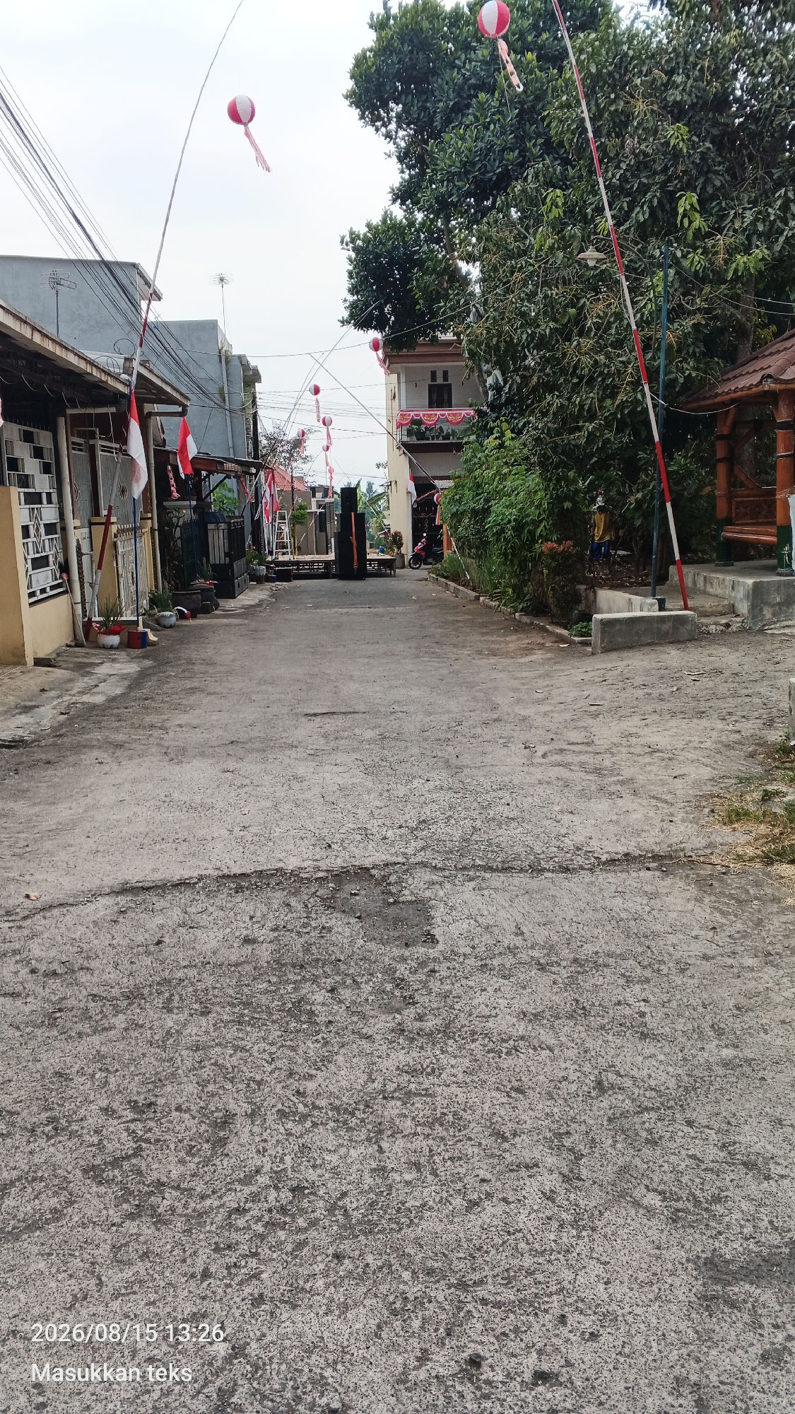 The image shows a narrow, paved street in a residential area of Jeru, Indonesia. The street is lined with houses and trees. Red and white decorations, including flags and balloons, are visible, suggesting a celebration or national holiday. There are also several speakers and a platform set up on the street, which might indicate an event is about to take place or has recently concluded. A motorcycle is parked on the side of the street. The sky is overcast, and the overall lighting is dim. A timestamp and text overlay are present at the bottom left of the image.