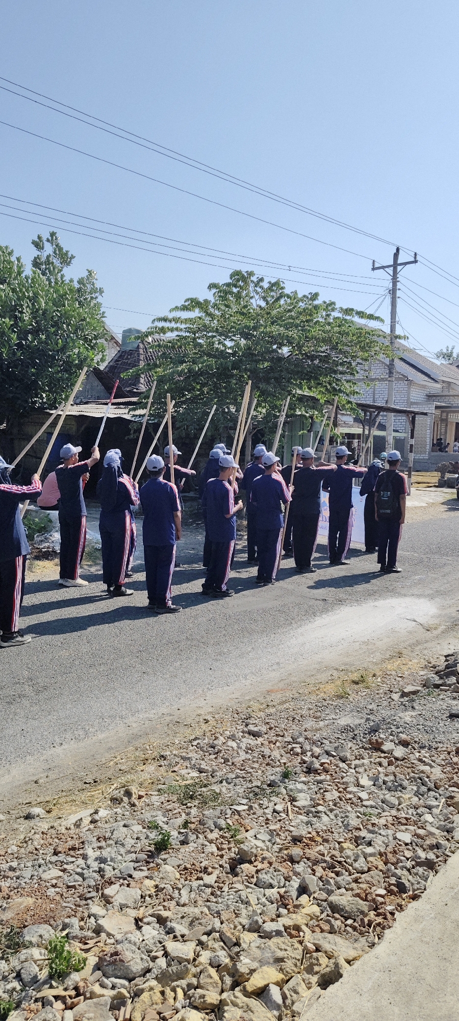 In Sukorejo, Indonesia, a group of individuals, likely students given their uniforms and age, stand in a line on an asphalt road. They are dressed in dark blue tracksuits with red and white stripes on the sides and matching blue caps. Each person holds a long wooden pole vertically, with the poles extending above their heads. The group is positioned on the right side of the road, facing away from the viewer. Behind them, houses with tiled roofs are visible, along with lush green trees and a utility pole with electrical wires. The ground on the side of the road is covered with gravel and scattered debris. The scene is illuminated by bright sunlight, casting shadows on the road.