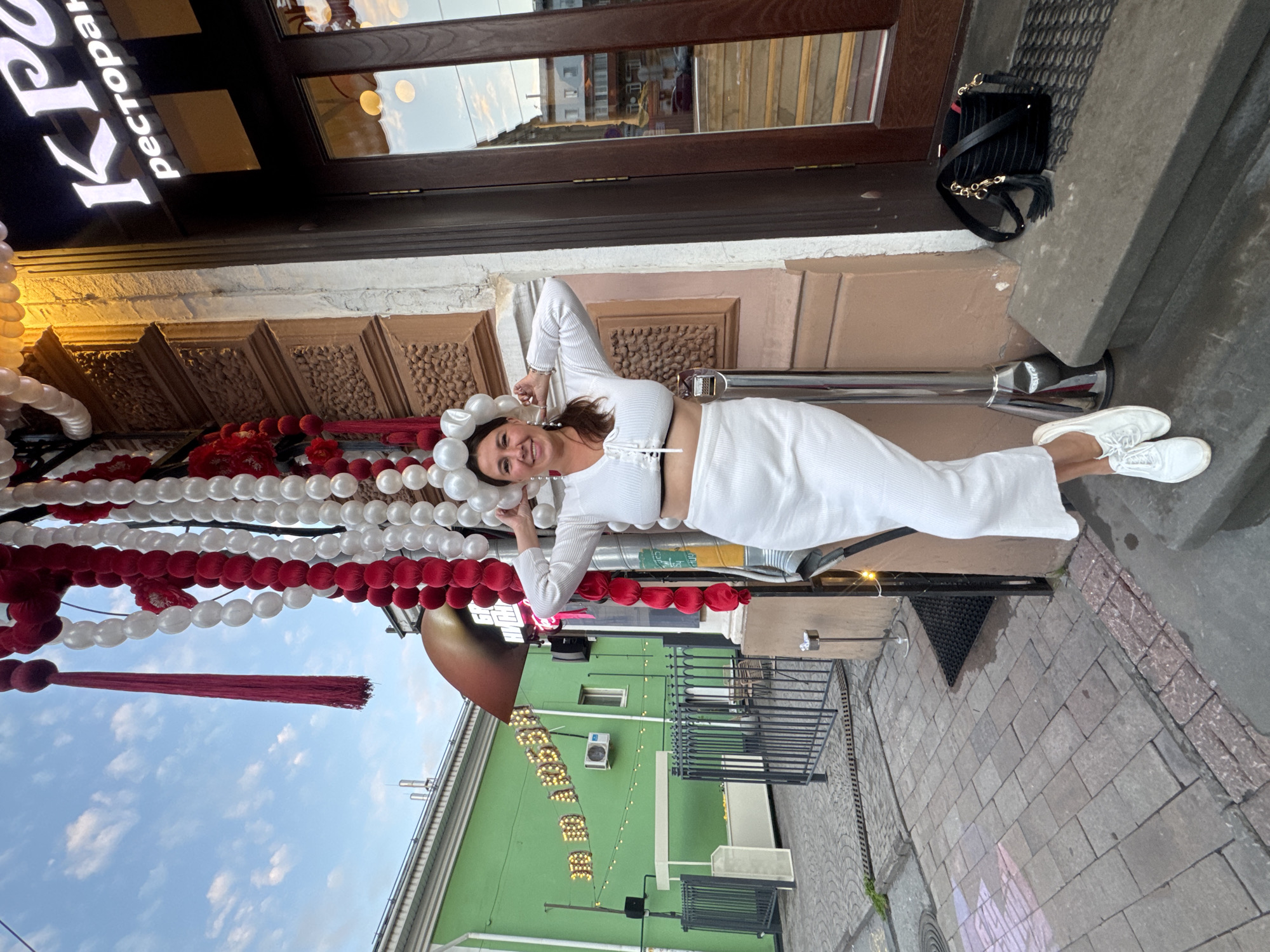 A woman in a white crop top and skirt stands outside a restaurant in Nizhny Novgorod, Russia, with a balloon archway behind her. The restaurant sign, "КР", is visible above the entrance. The woman is smiling and posing for the camera, holding her head with a balloon accessory. The scene takes place during the day, with a cloudy sky in the background.