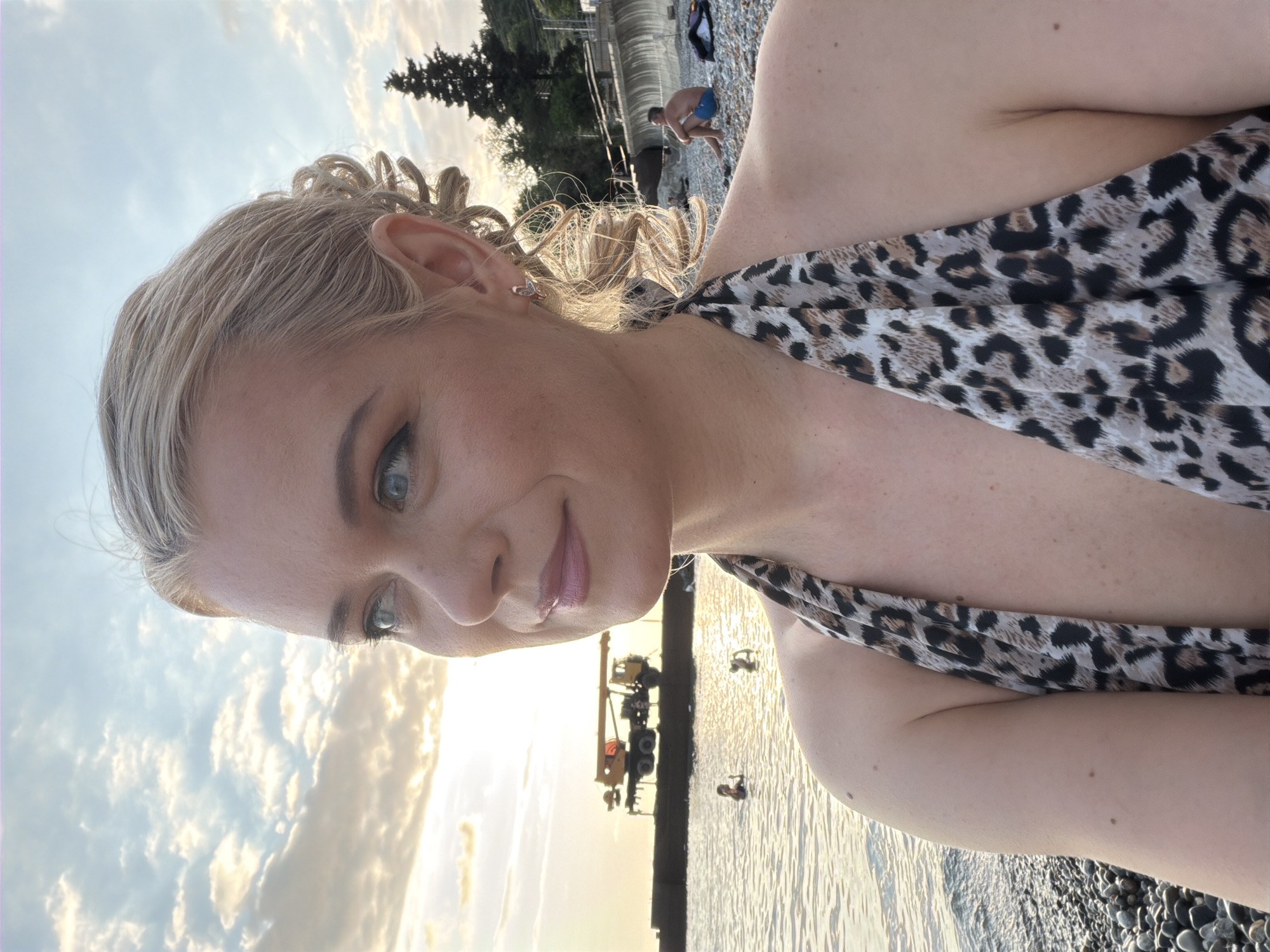 A medium shot captures a fair-skinned woman with blonde hair styled in an updo with some curls framing her face, looking to her left. She is wearing a leopard print halter top and is on a pebble beach, with the Black Sea behind her. The sun is setting, casting a warm glow on the scene. In the background, a yellow crane is visible on a pier, and a few people are in the water. Further back, a man sits on the beach, and trees line the coastline. The location is identified as Sochi, Russia.