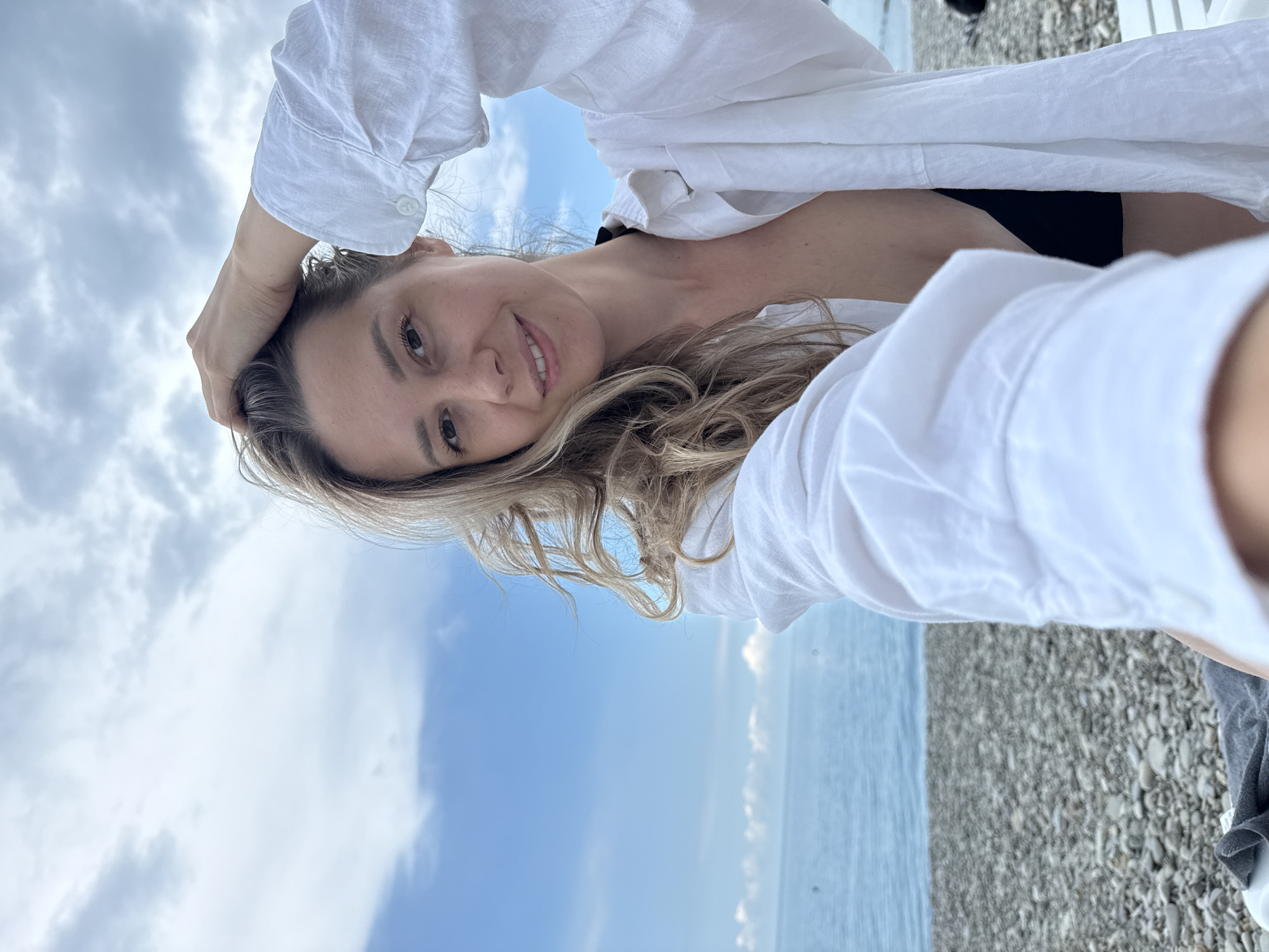 A woman with blonde, wavy hair and light skin is taking a selfie on a pebbled beach. She is wearing an unbuttoned white shirt over a black bikini top. The sky is cloudy and blue, and the sea is visible in the background. The location is the city of Nebug, Russia.