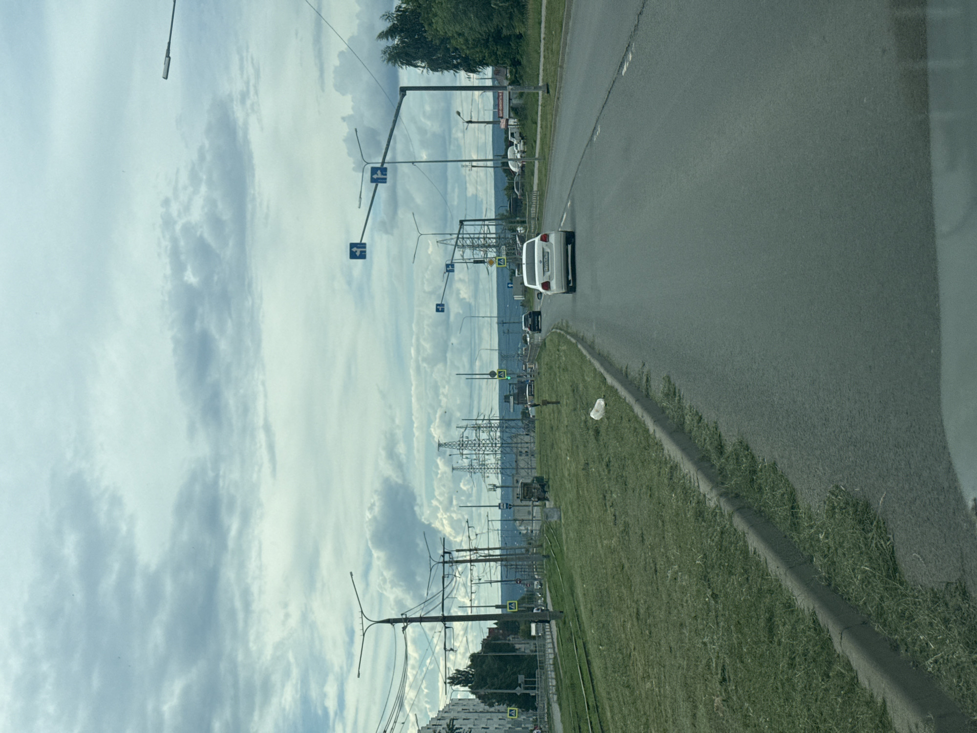 The image captures a view of a road in Samara, Russia, on a cloudy day. The road appears to be a multi-lane thoroughfare with a grassy median strip that has recently been mowed, with clippings visible along the curb. Several cars are on the road, including a white sedan in the foreground. Overhead, power lines and traffic lights are visible, along with tall electrical towers in the background. Buildings line the left side of the road, and trees are present on the right. The sky is filled with dramatic, grey clouds. There are no people clearly visible in the image, except for potentially distant figures.