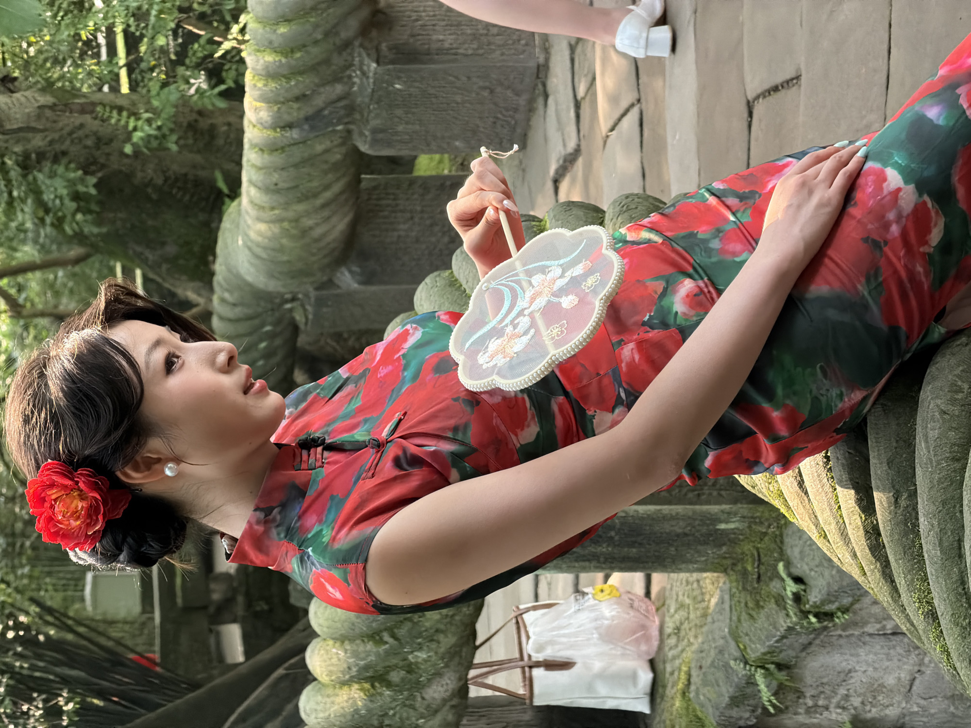 The scene depicts an outdoor setting in Yuzhong District, China, characterized by stone architectural elements and dense greenery. A woman, appearing to be of East Asian descent, is seated centrally on a low, weathered stone railing. Her dark hair is styled in an updo, adorned with a large, red multi-petaled flower behind her right ear. She wears white, spherical pearl stud earrings. Her attire is a short-sleeved cheongsam (qipao) with a high mandarin collar and frog button closure, patterned with large red and pink flowers and green leaves on a dark background.
The woman is leaning slightly forward, with her head turned to her right, and her gaze directed downwards. Her right hand holds a small, translucent white fan featuring light blue and peach-colored floral embroidery and a beaded edge. Her left hand rests on her lap, displaying light blue nail polish.
The stone railing she sits on is carved with a rope-like or spiral pattern and exhibits patches of green moss. In the background, other moss-covered stone structures and lush green foliage, including trees and plants, are visible. The ground to the right is paved with irregular stone slabs.
To the lower right, the lower leg and foot of another individual are partially visible, wearing a white shoe with a block heel. In the mid-ground on the left, behind the seated woman, a white tote bag with brown straps is placed on a stone surface. Adjacent to it is a clear plastic bag containing a pink liquid-filled object and a yellow cap. The scene is illuminated by natural daylight, casting distinct shadows and highlights.