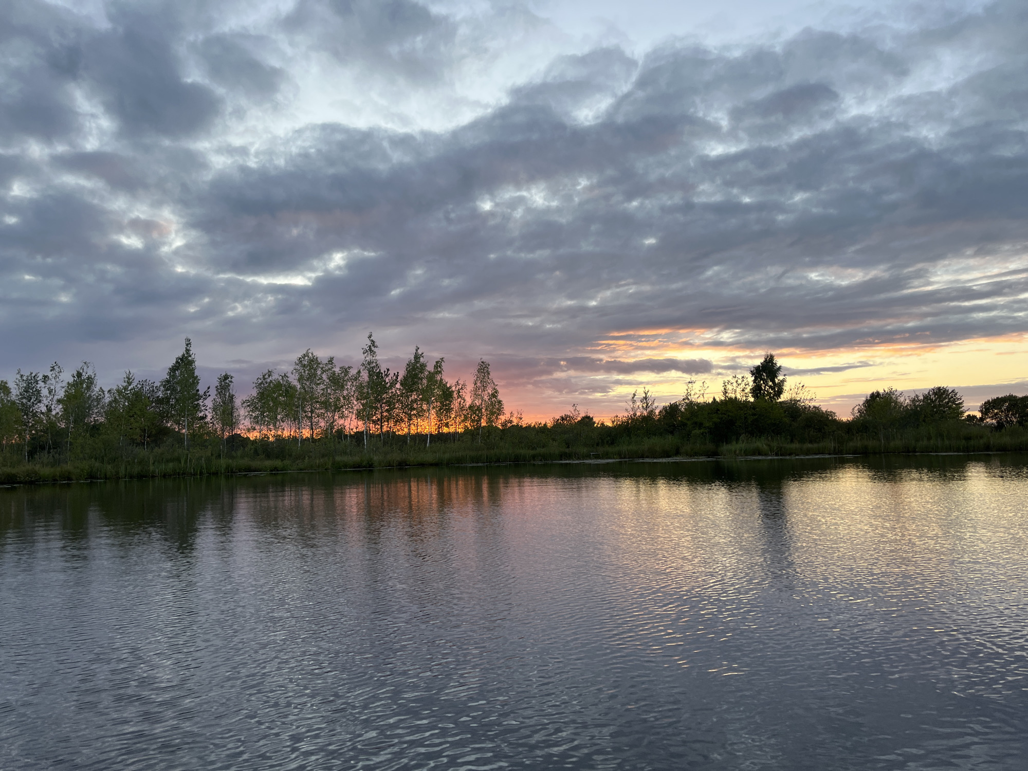 The image depicts a serene natural landscape at dusk, likely in or near Дунілавіцкі сельскі Савет, Belarus. A wide, calm body of water occupies the foreground, its surface rippling gently and reflecting the muted colors of the sky. The horizon is lined with a dense band of trees and vegetation, silhouetted against a sky transitioning from day to night. The clouds are a prominent feature, displaying various shades of gray, purple, and pink, with streaks of warm orange and yellow light from the setting sun breaking through in the distance. The reflections on the water mirror the colors of the sky and the dark outlines of the trees, creating a symmetrical and tranquil scene. No people or man-made structures are visible, emphasizing the natural and untouched quality of the environment. The overall impression is one of peacefulness and the beauty of twilight in a rural setting.