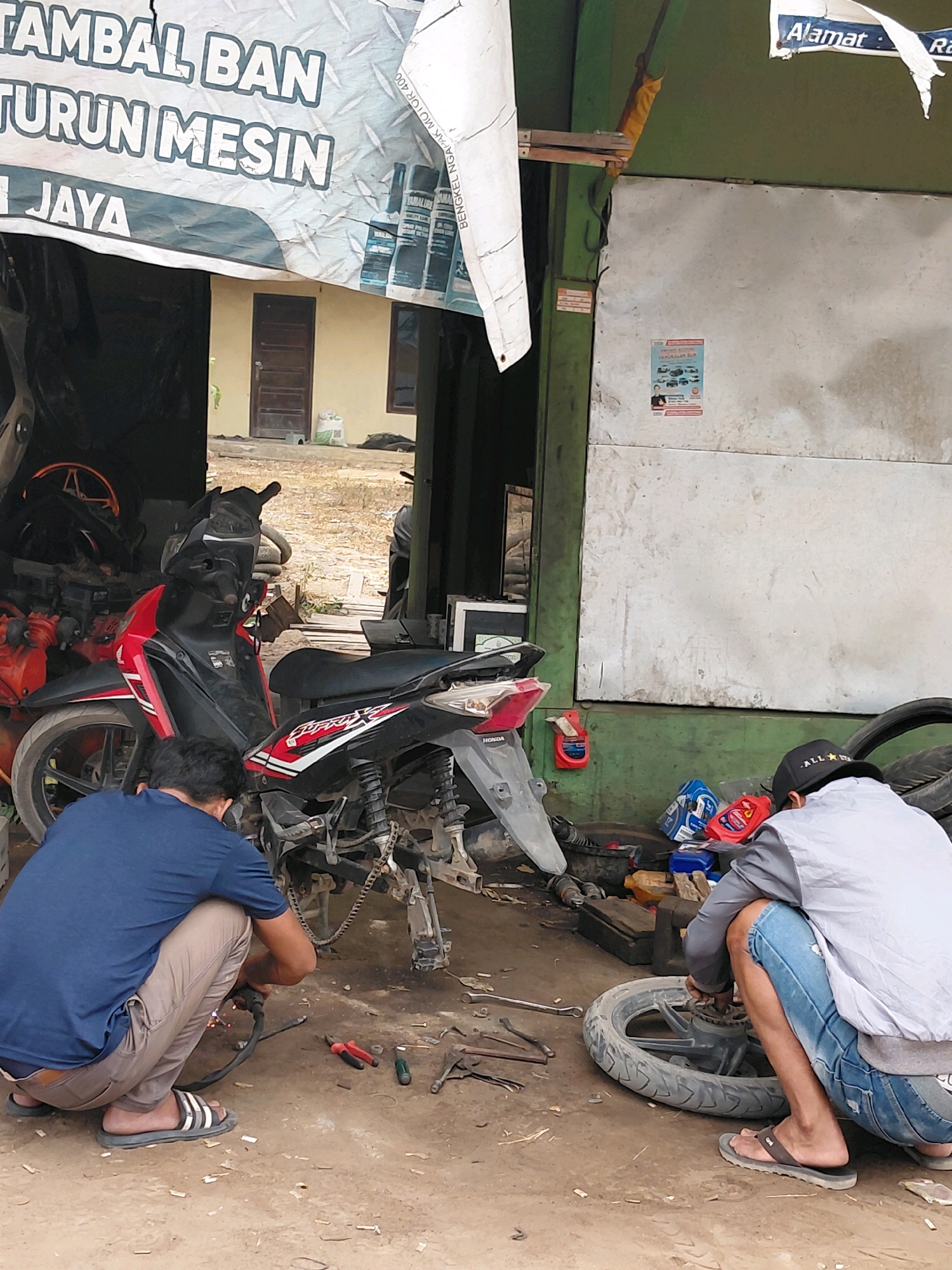 In Persiapan Mulya Raya, Indonesia, a mechanic shop is depicted with two individuals working on a motorcycle. The shop appears to be an open-air setup with a banner displaying "TAMBAL BAN TURUN MESIN JAYA" in large lettering. One mechanic, wearing a blue t-shirt and beige pants, is crouched down on the ground, appearing to be welding a part of the motorcycle with a welding torch. Tools like pliers and a screwdriver are scattered around him.
Another individual, wearing a gray jacket and blue jeans, is also crouched, working on the rear wheel of a motorcycle. He is focused on the wheel's components, with a tire lying next to him. Various bottles, likely containing oil or cleaning supplies, are visible near this mechanic. The ground is dirt and scattered with debris. The background shows a wall made of corrugated metal and a building with doors and windows. The scene captures the everyday operations of a local motorcycle repair service.