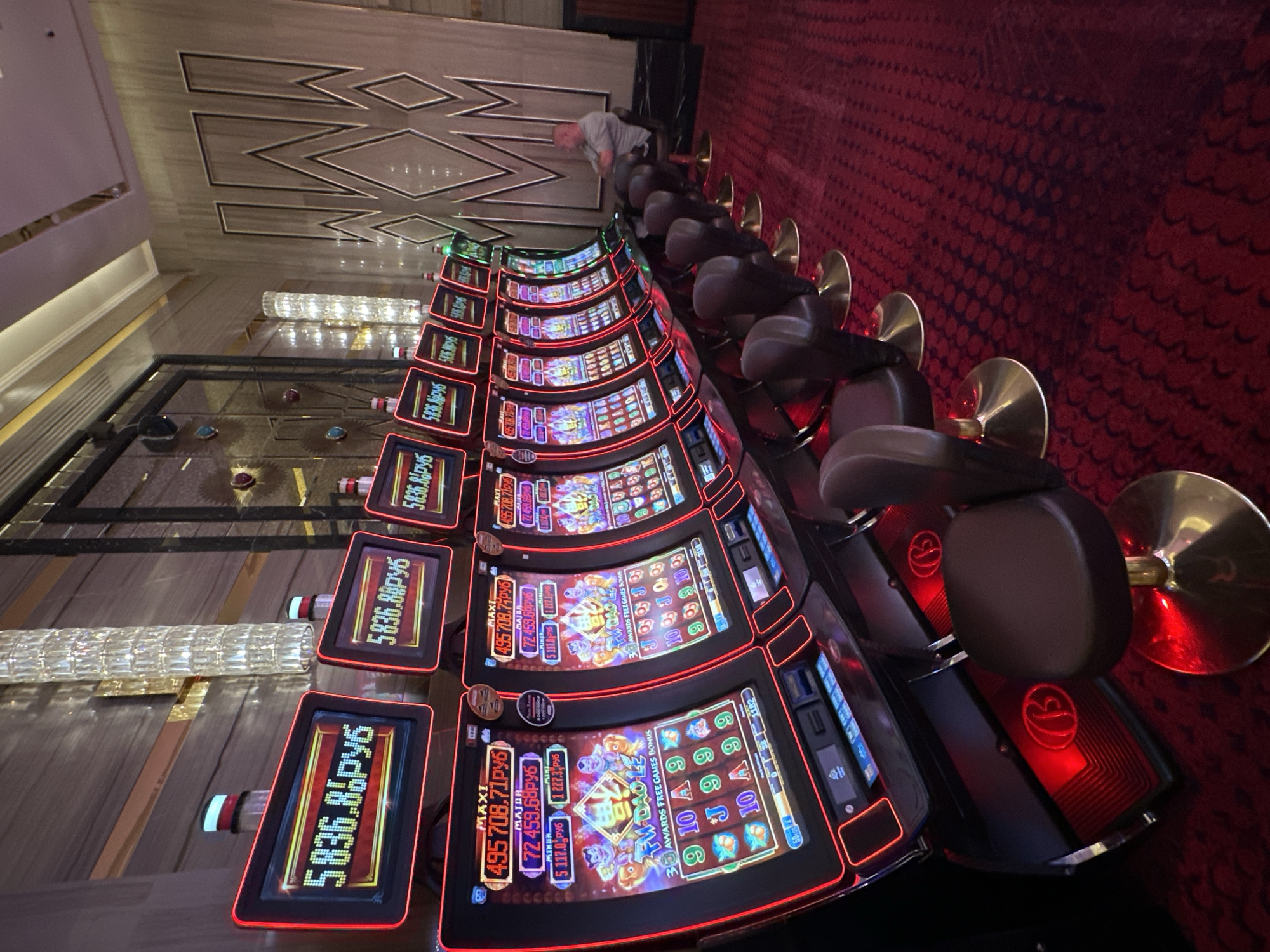 The image shows a row of slot machines in a casino, likely located in Estosadok, Russia. The slot machines have illuminated screens displaying amounts in Russian rubles (RUB) and game graphics. Some of the screens show progressive jackpot amounts, such as "495 708.71 RUB" and "72 459.68 RUB". There are several dark leather casino chairs in front of the machines, with one man sitting in a chair in the background, facing away from the camera. The casino floor is covered with a red carpet patterned with dark circular designs. The walls have a textured, reflective surface, and there are ornate crystal chandeliers hanging from the ceiling. The overall environment suggests a modern and well-lit casino setting.