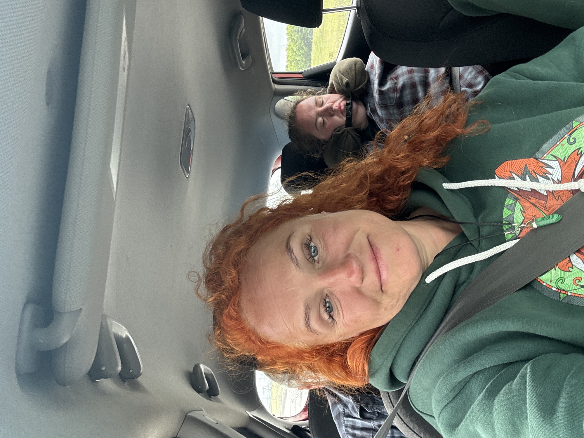 In the foreground of the image, a woman with vibrant red, curly hair is looking directly at the camera, smiling faintly. She is wearing a dark green hooded sweatshirt with a white drawstring and a colorful graphic design on the front. A seatbelt is visible across her chest. In the background, another person is sleeping in the back seat of a car, propped up by a travel pillow and appearing to be wearing a plaid shirt. The interior of the car is visible, with the ceiling and grab handles present. Outside the car window, a green, leafy landscape is visible under a cloudy sky. The location is stated to be Alnashi, Russia.