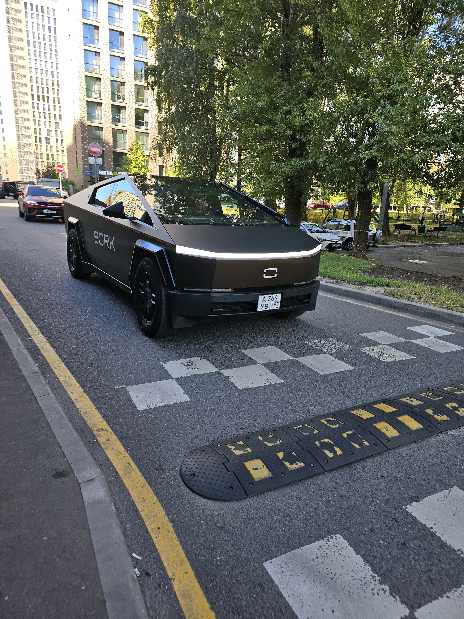 A black Tesla Cybertruck is parked on a city street in Khimki, Russia, near a pedestrian crossing with a speed bump. The truck has the word "BORK" printed on its side. A red car is parked behind the truck. In the background, a tall residential building and trees are visible. The sky is overcast.