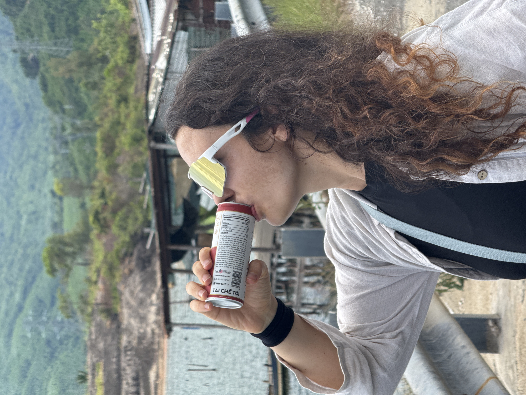 A medium shot captures a woman with curly brown hair wearing large, yellow-tinted sunglasses and a light-colored, open cardigan over a black top, drinking from a red and white can. She is positioned in the foreground, her face tilted upwards as she drinks. In the background, a blurred scene depicts lush green hills and some simple structures, indicative of a rural or semi-rural setting. The overall impression is one of a casual moment captured outdoors, potentially in a scenic area of Huế, Vietnam, characterized by natural landscapes and modest architecture.