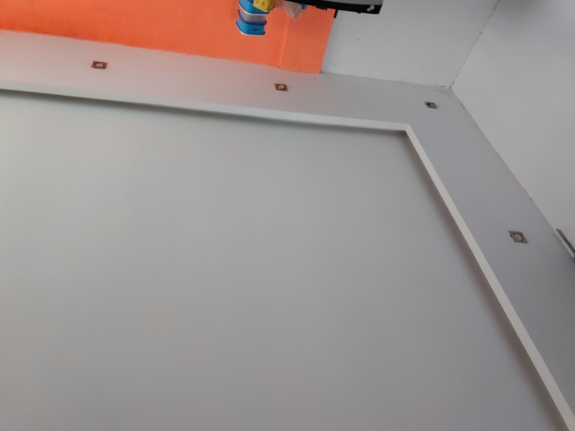 This image depicts an interior architectural space located in Santa Ana, Venezuela, showcasing a modern design with contrasting colors. The dominant feature is a large, smooth light gray wall, framed by a clean white border or trim. Above this gray section, a vibrant orange ceiling or wall portion extends across the upper part of the frame, providing a striking color block.
Several dark, small openings are visible, which appear to be recessed light fixtures or cutouts for utilities. One square opening is centrally positioned on the white trim beneath the orange area. Another circular fixture is located in the upper right corner on a lighter-colored wall. In the top-center, near the orange section, a distinctive object hangs, composed of stacked blue elements, possibly plastic containers or shades, topped with a yellow component. This could be a unique light fixture or decorative piece. The surfaces appear clean and well-maintained.
The image is a static shot of the room's features; no people are present, and no activity is occurring. The lighting is uniform, characteristic of an indoor setting. There is no visible text anywhere in the frame.