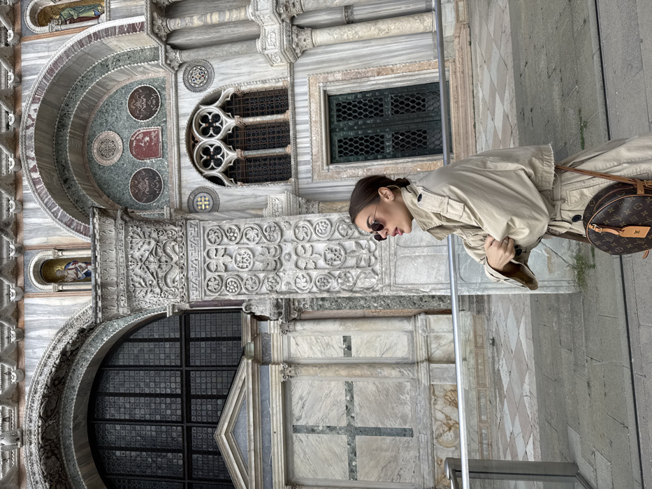A female individual stands in front of the ornate facade of St. Mark's Basilica in Venice, Italy. She is positioned in the mid-ground, facing left with her head tilted slightly upward and her mouth partially open. She wears dark, round sunglasses and a beige trench coat, belted at the waist, with the sleeves pushed up. Her dark hair is styled in a bun. A brown circular Louis Vuitton monogram handbag with a luggage tag is slung over her right shoulder. Her hands are clasped in front of her, resting on a foreground railing. The basilica facade behind her displays intricate Byzantine-style architecture featuring marble columns, arches, detailed carvings, and mosaic elements in shades of grey, white, green, and red. Notable architectural features include a large arched window with a grid pattern on the left, an ornate column with relief carvings, a gothic-style window with quatrefoil tracery, and a dark green double door with a grid pattern. Niches containing small figures are visible above the arches. The ground consists of patterned grey and white flagstones.