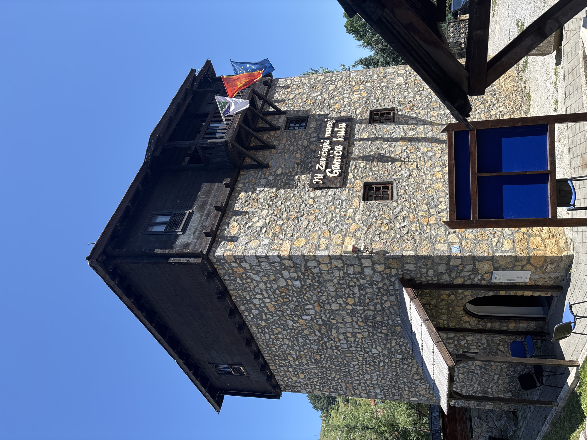 A stone building with a dark wooden upper story stands against a clear blue sky. The building has a single entrance under a sloped wooden awning. Three chairs, two blue and one black, are placed on the paved walkway in front of the entrance. A wooden sign with white lettering is mounted on the stone wall, reading "JU Zavičajni muzej Ganića kula". Three flags are displayed on a balcony above the sign. In the foreground, a wooden display board with two blue panels is positioned on the walkway. The scene is illuminated by bright sunlight, casting shadows from the building and the display board.