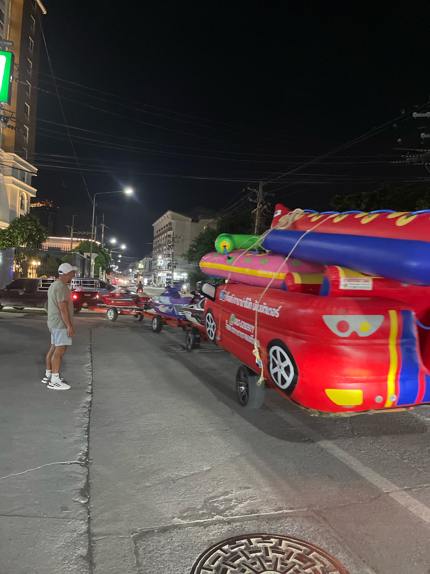 A man wearing a light brown t-shirt, light blue shorts, and white athletic shoes with black socks stands on a paved sidewalk facing away from the camera. He is looking at a bright red inflatable water slide on a trailer. The trailer is attached to a small car, and it is loaded with additional inflatable water toys. Behind the trailer, several jet skis are visible on separate trailers, also in the street. The scene takes place at night on a street in Pattaya City, Thailand, with streetlights illuminating the area. Buildings and power lines are visible in the background.