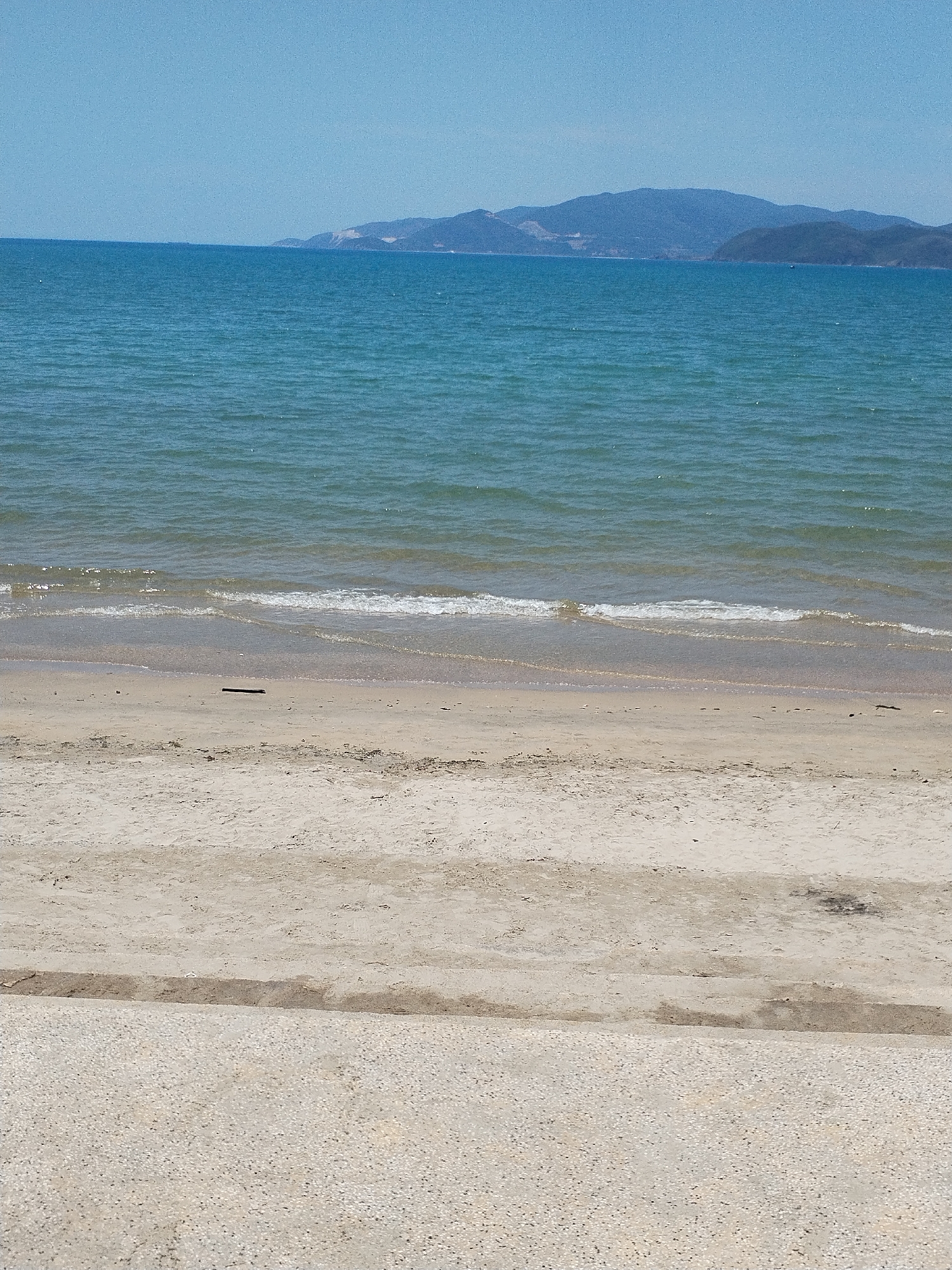 The image depicts a beach in Nha Trang Ward, Vietnam. The foreground consists of a light gray concrete surface, transitioning to a sandy beach with varying levels of sand compaction. The ocean occupies the midground, with gentle waves reaching the shore. In the background, there are hills or mountains visible against a clear blue sky. There are no people visible in the image. A small dark object is present on the sandy beach.