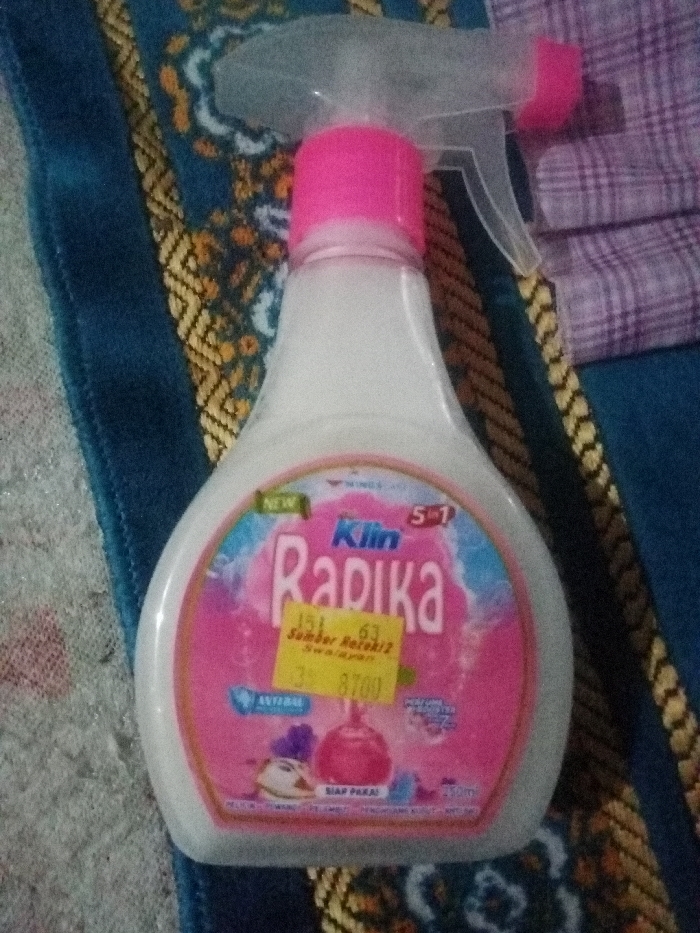 A bottle of "Klin Radika" cleaner is displayed on a patterned surface, possibly fabric. The bottle has a pink and white color scheme, with a clear spray nozzle and a pink cap. A yellow price tag is affixed to the front of the bottle. The cleaner is labeled as "5 in 1" and appears to be a household cleaning product. A purple plaid fabric is visible in the background on the upper right.