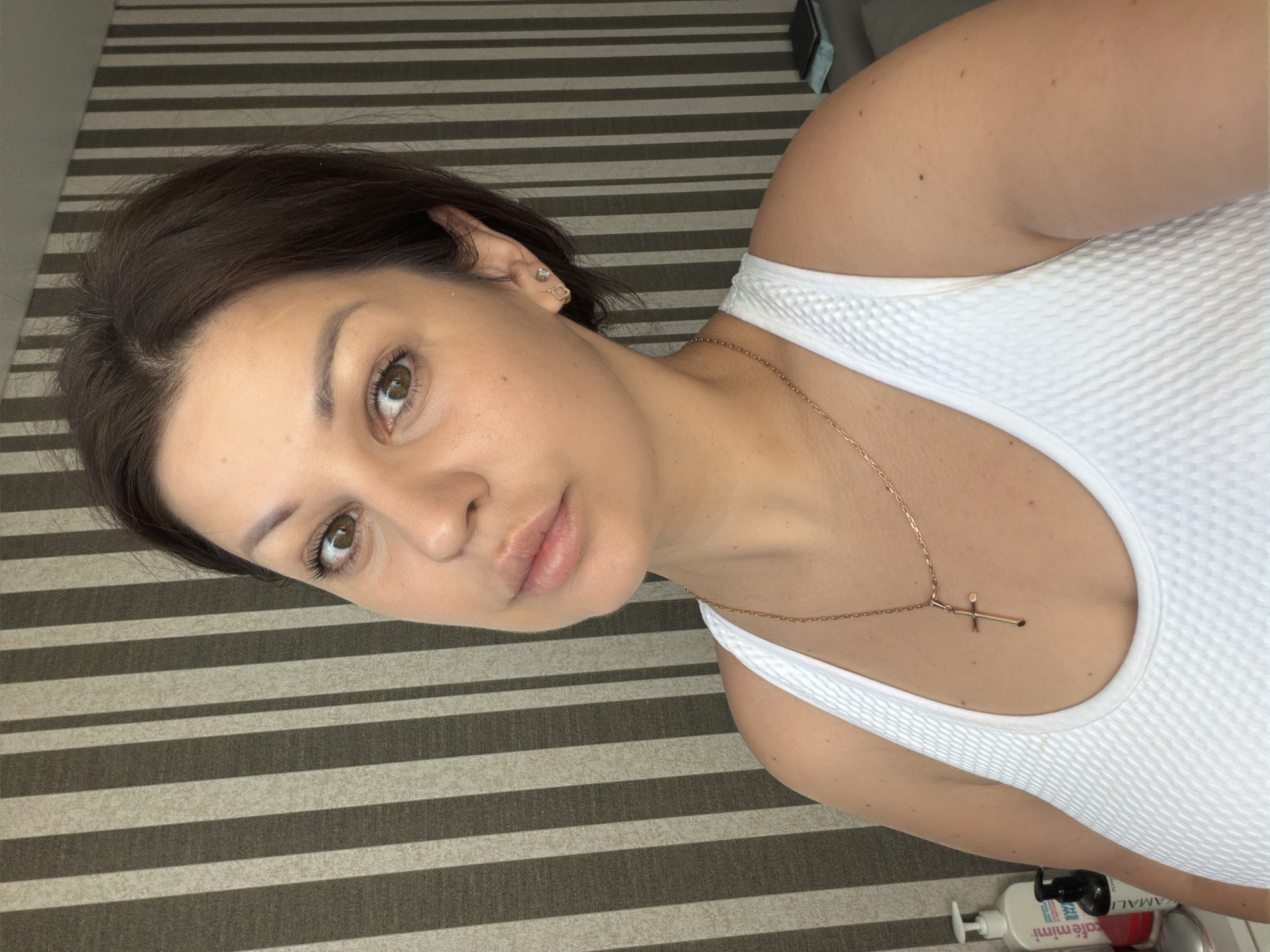 The image shows a medium shot of a woman with brown hair and brown eyes. She is wearing a white tank top and a gold cross necklace. She has on light makeup and a subtle smile. She is looking directly at the camera. The background is a striped wall with alternating dark brown and light beige stripes. To the left of the woman, there are two bottles of what appear to be cosmetic products on a surface. The scene appears to be indoors, possibly in a home or hotel room. There is no indication of the city of Sochi, Russia in the image itself.