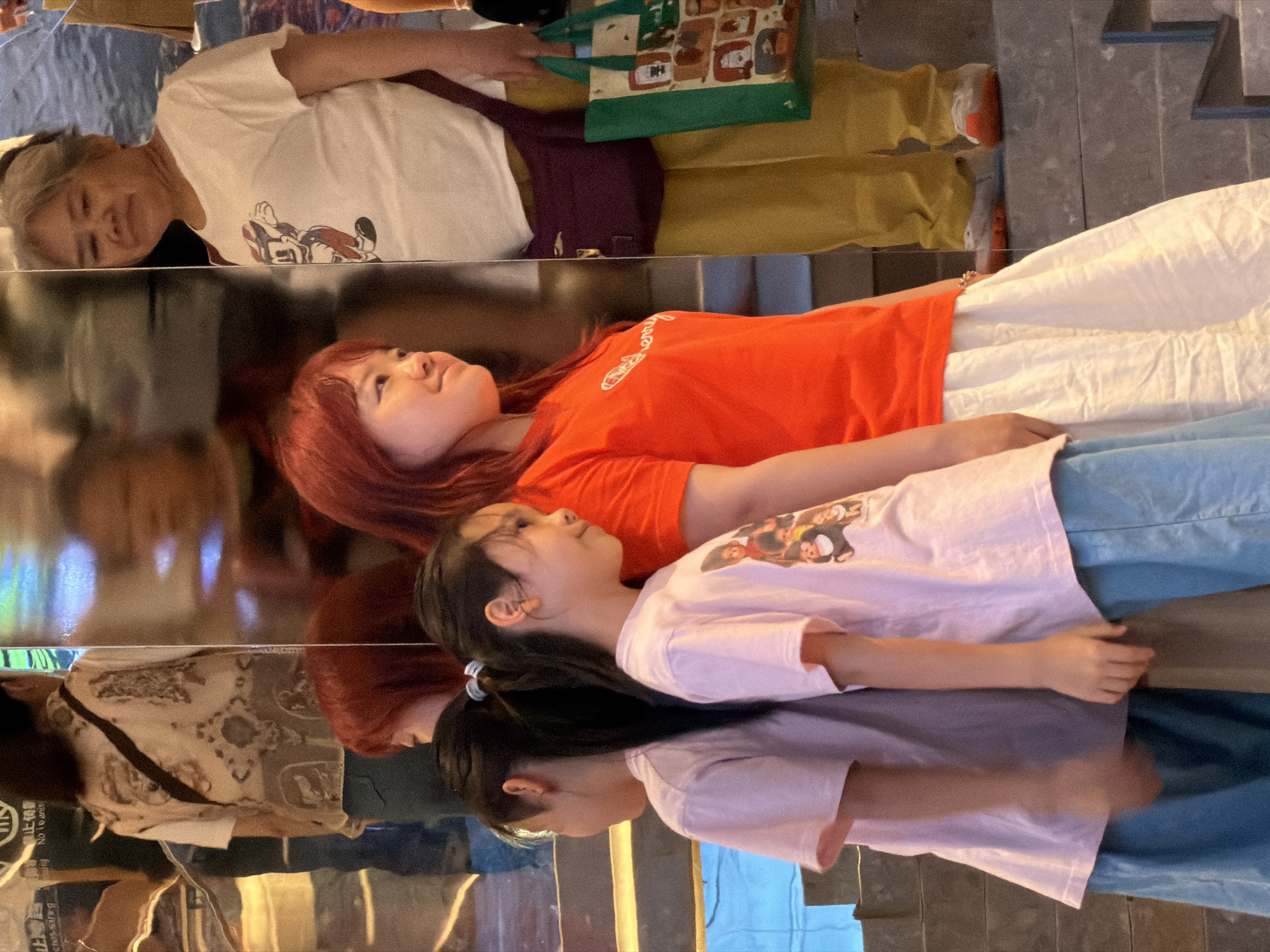 The image depicts a scene in the Yuzhong District of Chongqing, China, featuring a group of people reflected in a large, mirrored surface. In the foreground, two young girls stand side-by-side, their backs partially turned to the viewer. They both wear light pink t-shirts and light blue pants. To their right, a young woman with reddish hair wears a bright orange t-shirt and a white skirt. Behind her, an older woman in a white t-shirt with a cartoon graphic stands with a purple fanny pack and holds a green tote bag. The mirrored surface reflects multiple figures and the surrounding environment, which includes stairs and an indication of an indoor setting with some lighting. The overall composition is a reflection-heavy snapshot of a moment in a public or commercial space within the Yuzhong District.