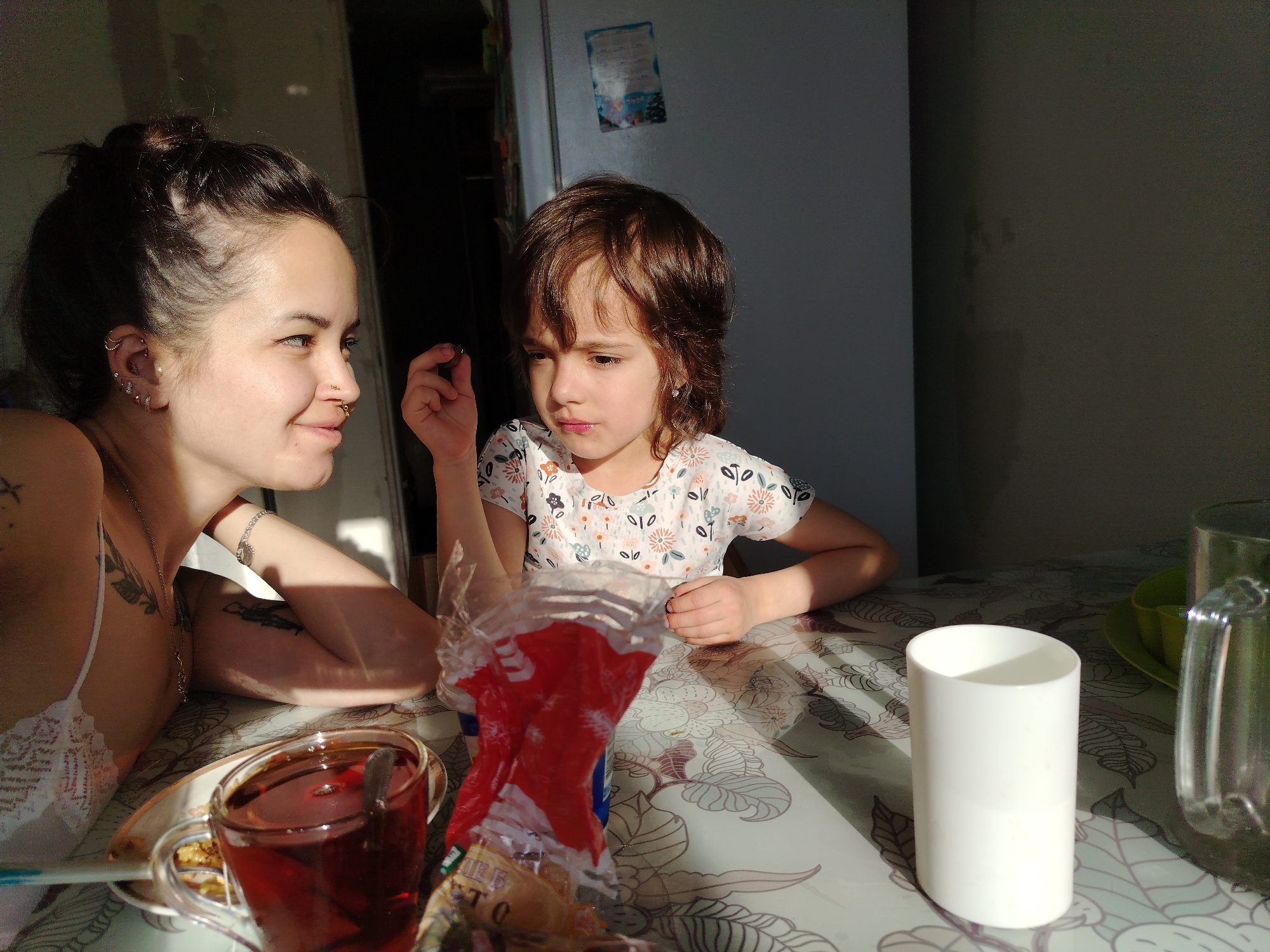 The image depicts two individuals, an adult female and a young girl, sitting at a table in what appears to be a kitchen or dining area. The adult female, positioned on the left side of the frame, has dark hair pulled back in a bun, multiple piercings on her ear and nose, and tattoos visible on her arm and chest. She is wearing a light-colored top and is looking towards the girl. The young girl, on the right side of the frame, has short brown hair and is wearing a printed short-sleeved shirt. She is holding something small in her hand and appears to be deep in thought.
On the table, there is a cup of tea, a plate with food, a crumpled package, a white cylindrical object (possibly a cup or container), and a glass pitcher. The table has a patterned tablecloth. The background reveals a refrigerator and a plain wall. The lighting suggests the presence of sunlight entering from a window.
There is no specific information in the image or the instructions indicating the location is Noviy, Russia, but the overall impression is that this is a domestic, possibly Eastern European, scene.
