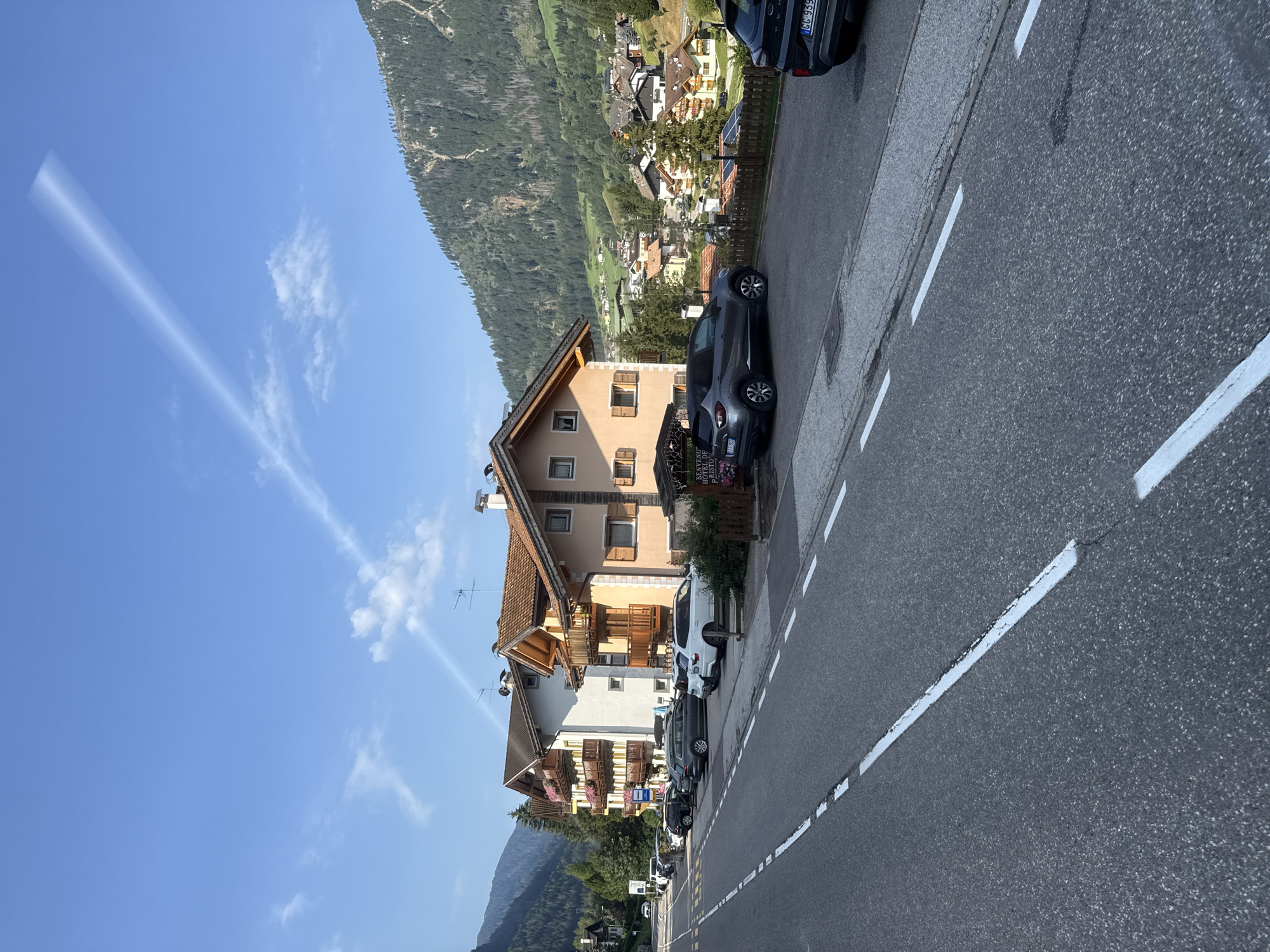 The image shows a street in Moena, Italy, with several buildings and cars parked along the side of the road. The buildings are typical of a mountain town, with sloped roofs and wooden balconies. The mountains in the background are covered in trees. The sky is blue with some clouds. There are no people visible in the image.