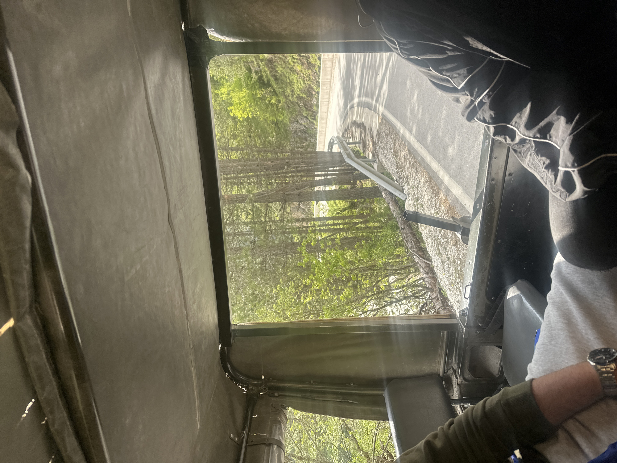 The image depicts a view from inside a military vehicle, likely a Humvee, with a focus on the passenger-side window. Outside the window, a road with a guardrail is visible, lined by trees with green foliage, suggesting springtime. The road curves slightly. Inside the vehicle, part of the roof and side panel are visible, showing the canvas and metal frame construction. A person is partially visible in the passenger seat, wearing a grey shirt, a black jacket with white trim, and a watch on their left wrist. The environment appears to be in a forested area. There is no information from which to determine the specific location in Iagodina, Bulgaria.