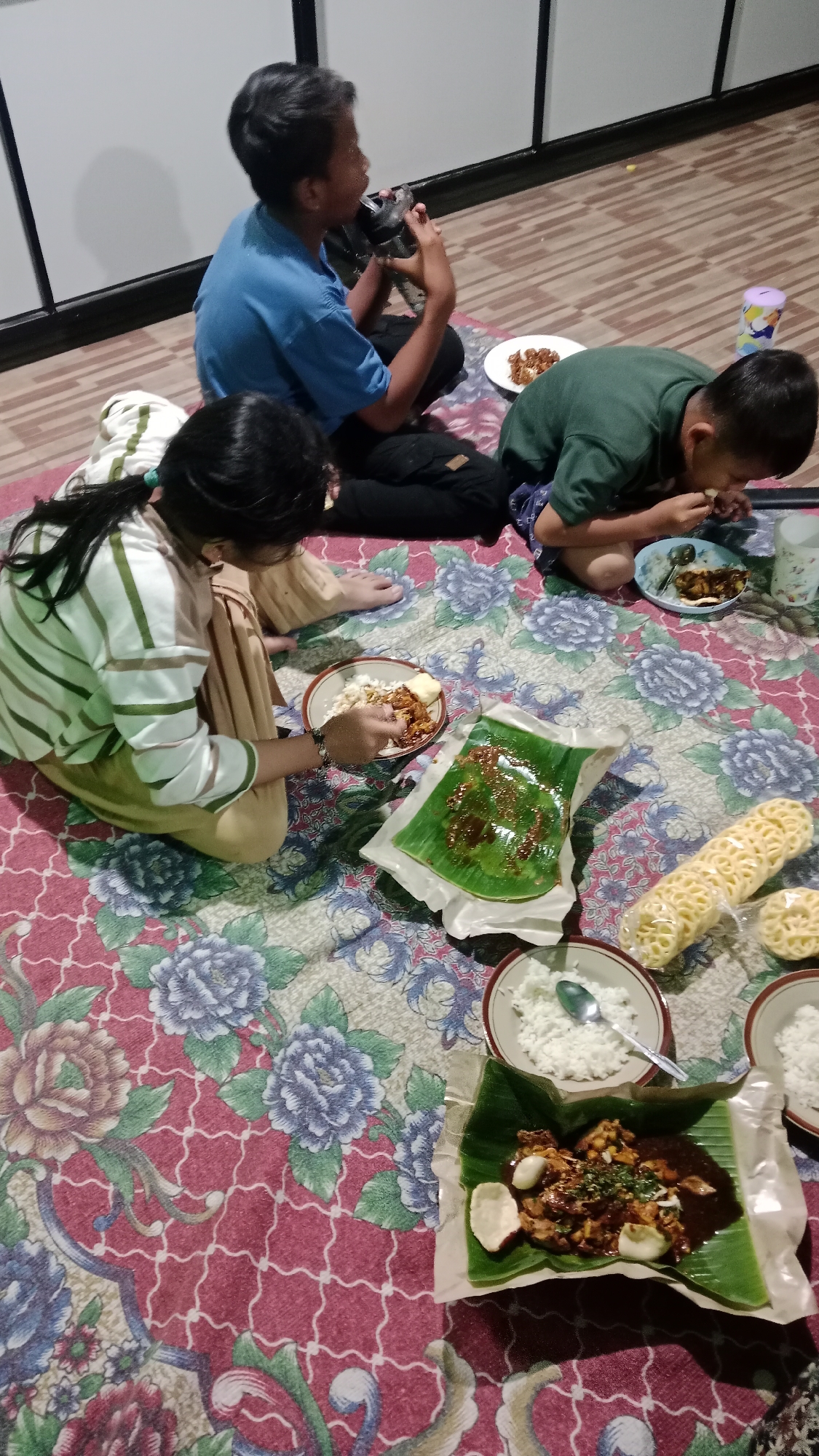 The image depicts three children sitting on a floral-patterned mat on the floor, likely inside a home. They are engaged in eating a meal served on banana leaves and in bowls. One child, wearing a blue t-shirt, is holding a black object to their mouth, possibly a camera or phone. Another child, in a green shirt, is eating from a plate. A third child, with dark hair and wearing a striped shirt, is also eating from a bowl. Several plates of rice, food wrapped in banana leaves, and crackers are visible. The environment appears to be a residential interior with a wooden floor visible in the background. The lighting suggests it is evening or indoors. The presence of Indonesian food and the style of the home decor are consistent with a scene in Madiun, Indonesia.