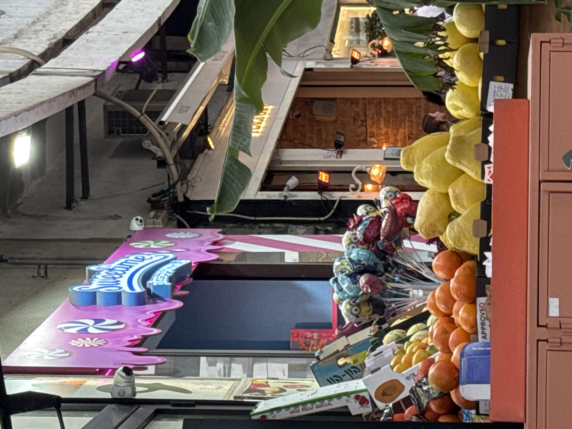 The image depicts a view in Tel-Aviv, Israel, looking upwards at a street scene with various elements clustered together. A "Sweettime" sign, designed to resemble melting candy with blue and white lettering, is visible on the left. Above and behind the sign, there are building facades, including windows, exposed piping, and a lighting fixture. To the right, a collection of objects is displayed, including balloons, oranges, lemons, and packaged goods. A narrow, vertically-oriented piece of red furniture (possibly a locker) is on the extreme right. The scene is cluttered and colorful, with a mix of architectural details and commercial displays.
