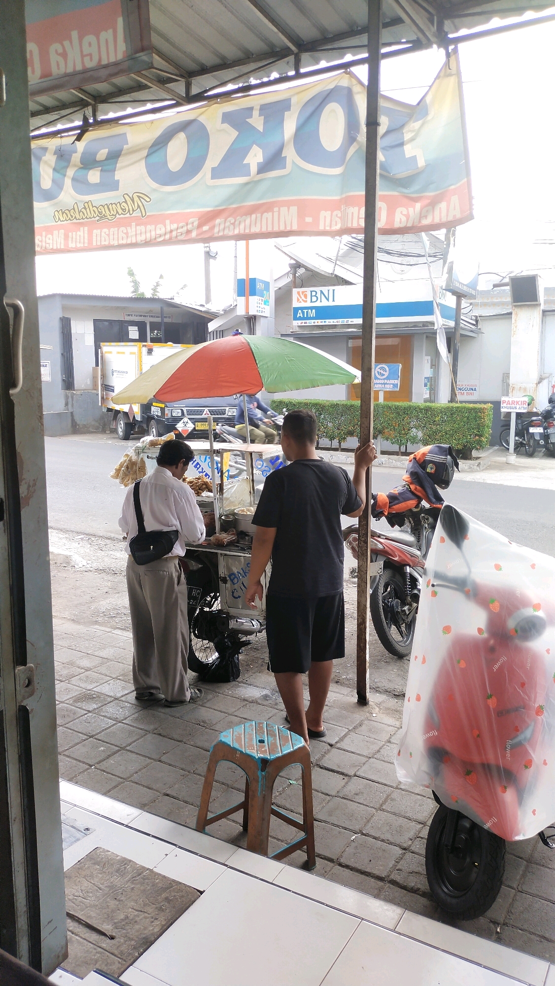 The image captures a street scene in Jember, Indonesia, likely outside a shop. In the foreground, a food cart is visible, with two men standing near it. One man, wearing a white shirt and grey pants, appears to be attending to the cart. The other man, in a black t-shirt and shorts, is facing away from the camera. Behind them, a red motorcycle covered with a plastic sheet decorated with strawberries is parked. A small, blue and brown stool is placed on the sidewalk. The background shows a street with vehicles, buildings, and a BNI ATM sign. A large banner with blue and yellow lettering is hanging overhead. The overall impression is a snapshot of daily life in a commercial area.