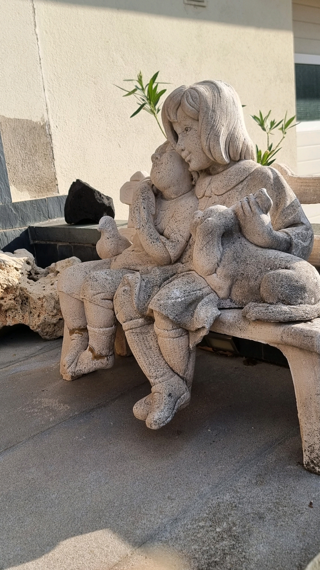 In the city of Sant Joan Les Fonts, Spain, a stone sculpture depicts two children and a dog sitting on a bench. The sculpture is positioned outdoors, with a textured beige wall in the background and a light-colored concrete surface in the foreground. Green foliage is visible behind the sculpture. The larger child, a girl with short hair, is embracing a smaller child, who is nestled against her. A dog sculpture is seated to the right of the children. The stone material of the sculpture shows signs of weathering and some discoloration. The scene is illuminated by natural sunlight, casting shadows on the ground.