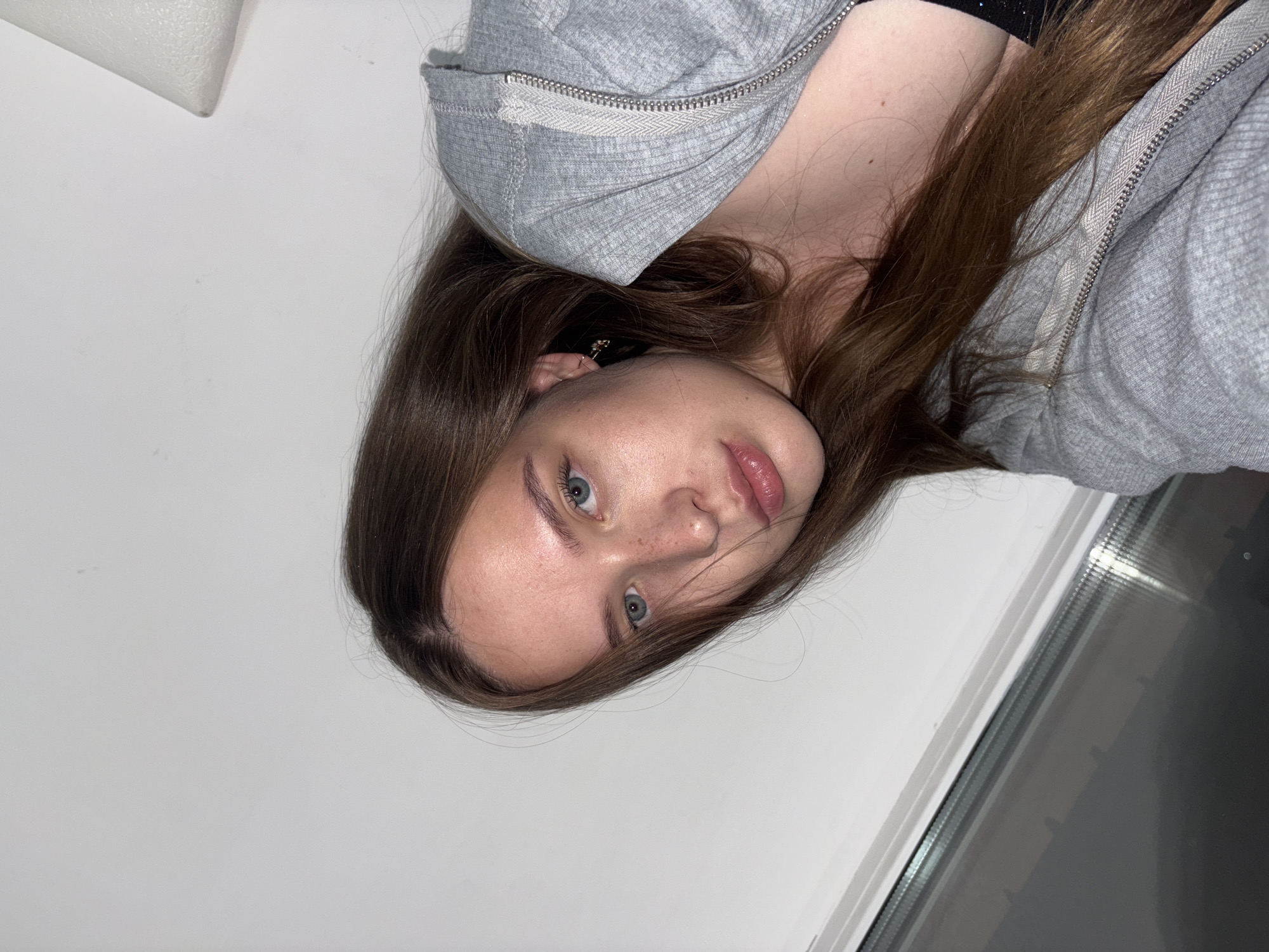 A young Caucasian woman with long, brown hair and blue eyes is captured in a close-up, slightly angled shot against a white wall. She is wearing a grey zip-up hoodie and a black top. Her head is tilted, and she is looking towards the camera. The background includes a window with a view of what appears to be a building across the street, suggesting an urban setting. The lighting is soft, possibly from a window or indoor lamp. The scene seems to be indoors, in what could be a residential apartment or a studio, with no specific identifying markers of Chaikovsky, Russia visible.