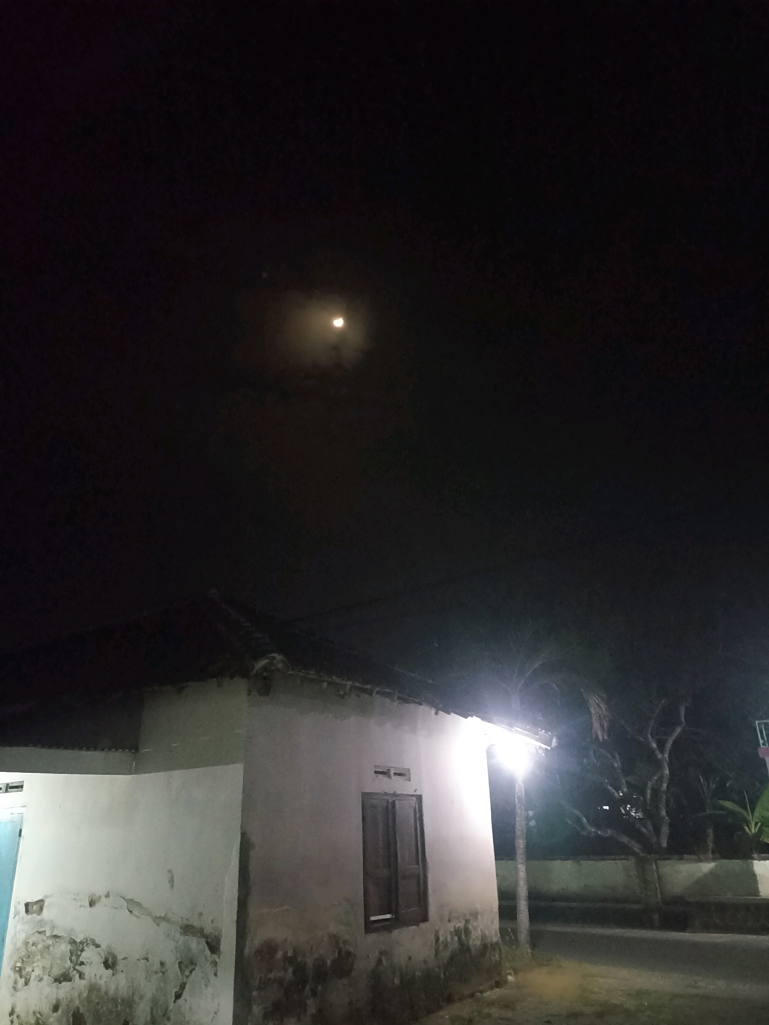This is a vertical, low-light photograph taken at night in the city of Sumberharjo, Indonesia. The image shows a partially lit, dilapidated building with a dark, corrugated roof. A single window with wooden shutters is visible on the side of the building. To the right of the building, a bright, modern street lamp illuminates the scene, casting strong shadows. In the background, bare tree branches are silhouetted against the dark sky. The moon, appearing as a crescent, is faintly visible through a thin layer of clouds in the upper portion of the frame. The ground in front of the building appears to be a dirt road or pathway.