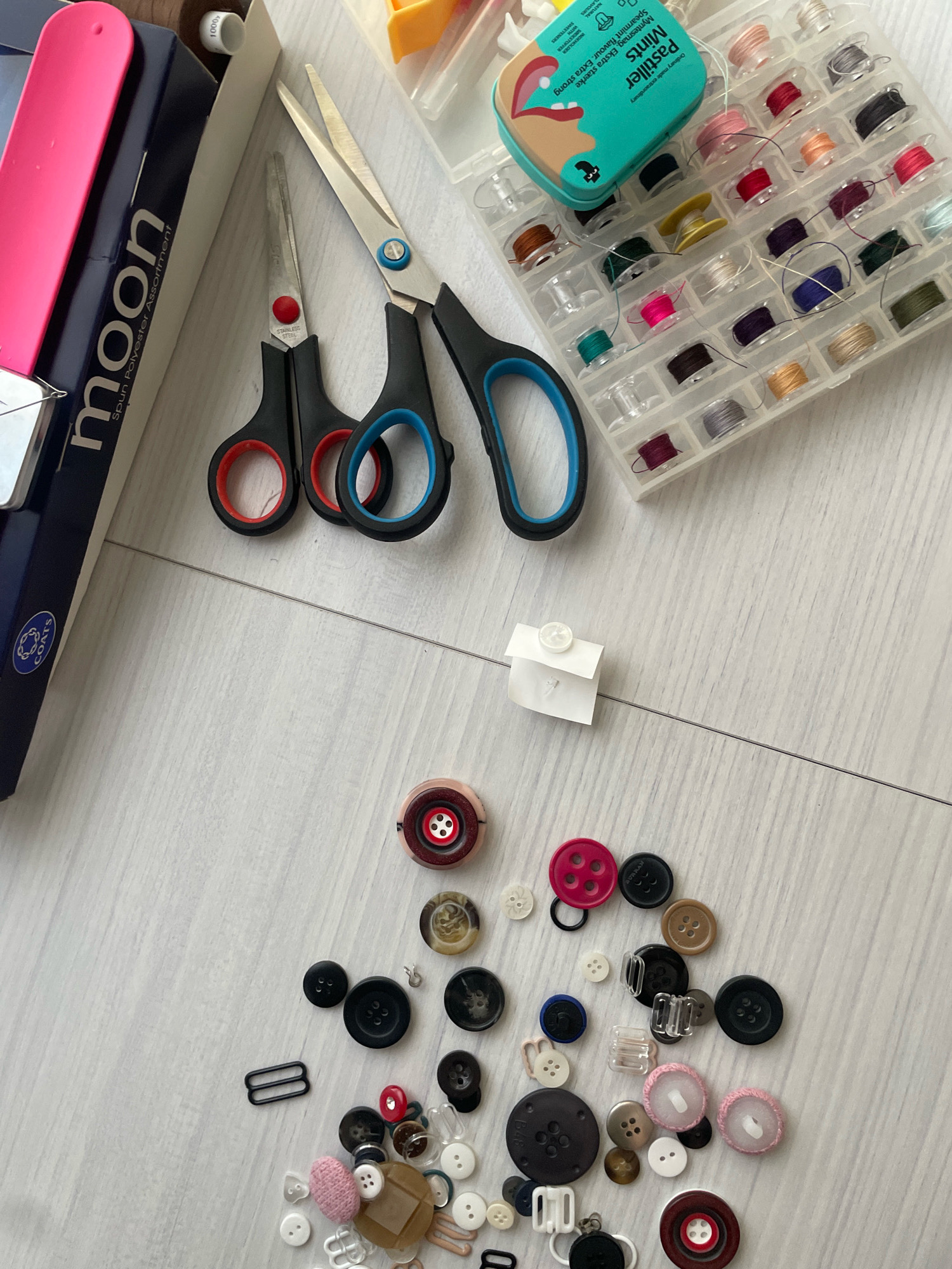 The image shows a collection of sewing supplies laid out on a light gray, wooden surface. The items include scissors, a box of thread spools, a tin of pastilles, and a variety of buttons. Specifically, there are two pairs of scissors, one with red handles and the other with black and blue handles. The thread spool box contains numerous spools of colored thread. Scattered across the surface are many buttons of different sizes, colors, and materials. Some buttons are plain, while others have decorative patterns or multiple holes. There is also a small white folded piece of paper with a white button attached to it by a thread. The lighting suggests an indoor setting. The image is a flat lay, taken from directly above. There are no people visible in the image. The location is Atarfe, Spain.