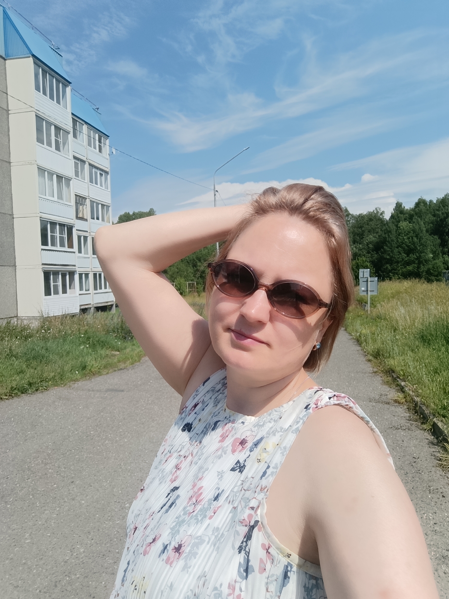 A light-skinned woman in her 30s or 40s, with shoulder-length blonde hair, is taking a selfie outdoors on a sunny day. She is wearing sunglasses and a white dress with a floral pattern. Her left arm is raised, and her hand is resting on her head. The background shows a paved path, a grassy area with trees in the distance, and a multi-story apartment building with a blue roof. A signpost is visible in the background. The scene appears to be in an urban or suburban area of Markovsky, Russia.