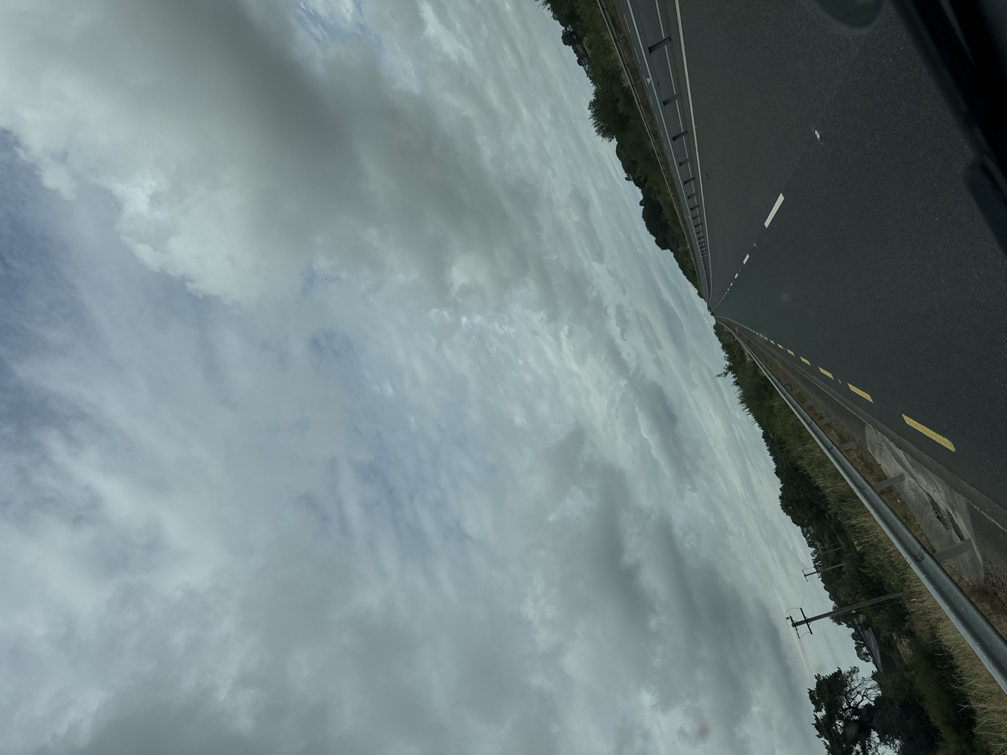 The image shows a view from inside a vehicle traveling on a highway. The road is dark asphalt with white lane markings and yellow solid lines on the side. There is a metal guardrail along the left side of the road, separating it from a grassy embankment with some trees and vegetation. In the distance, more trees and rolling hills are visible under a cloudy sky. Two utility poles are visible on the left side of the road. The sky is filled with various shades of gray clouds, indicating an overcast day. The perspective suggests the vehicle is moving forward, away from the viewer. The location is likely a rural area within the New Ross Municipal District of Ireland, given the typical road infrastructure and landscape. There are no people or other vehicles visible in the frame.