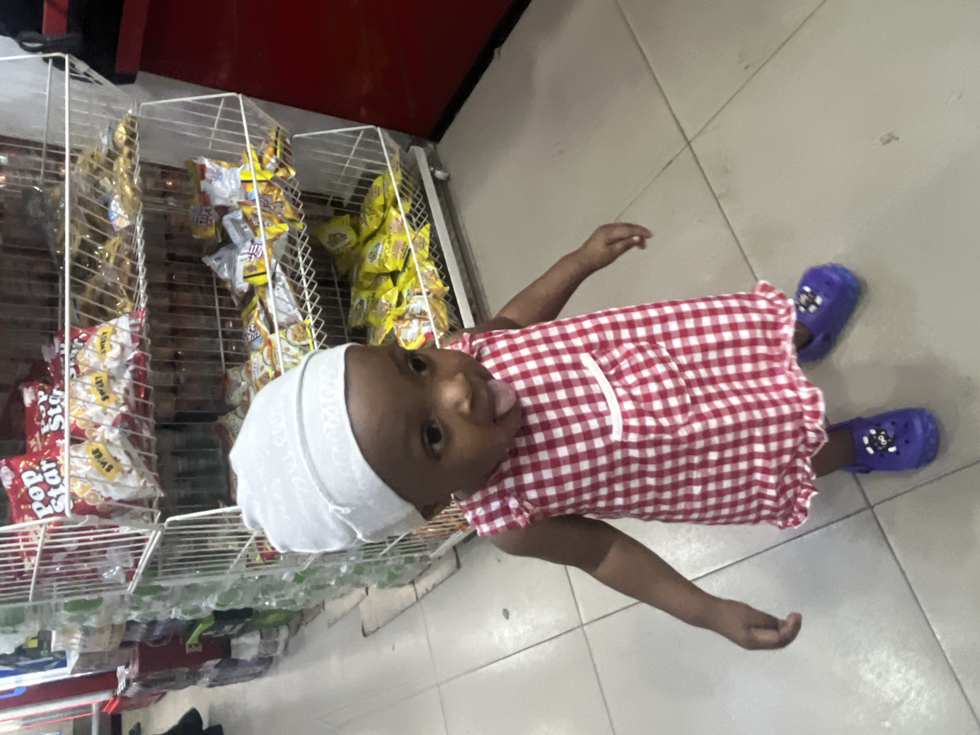 A young child stands in an Ado, Nigeria store. The child is wearing a white head covering, a red and white checkered dress, and purple Crocs. The child is sticking out their tongue. Shelves filled with various food items are visible next to the child.
