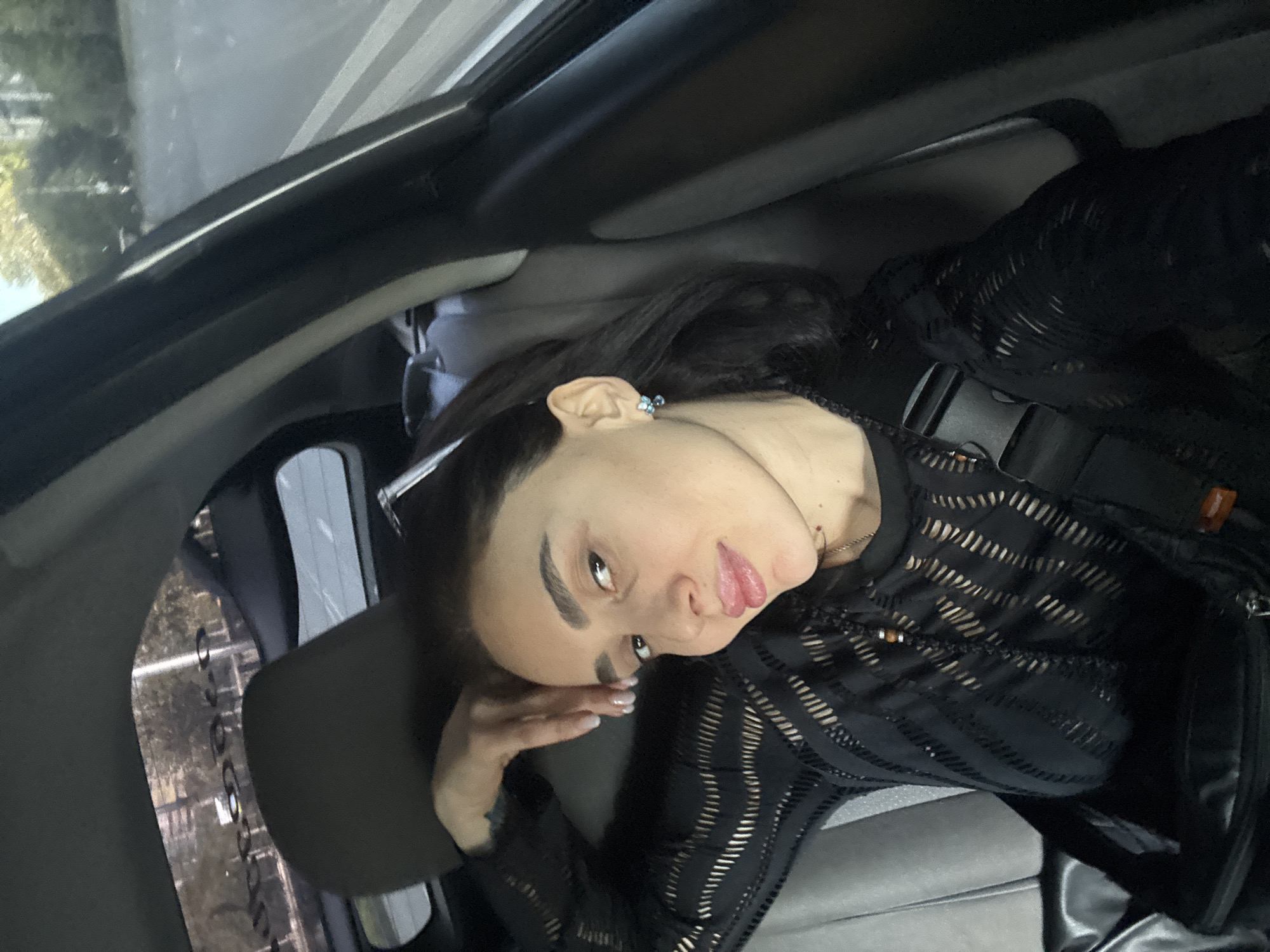 A person with dark hair and light skin is inside a car, possibly in Tbilisi, Georgia, judging by the blurred view outside the window. The person is wearing a black, semi-transparent top and has their hand resting on their head. The car's interior is visible, including the roof, seats, and window frame. Outside, there are street lights and buildings, suggesting an urban environment.
