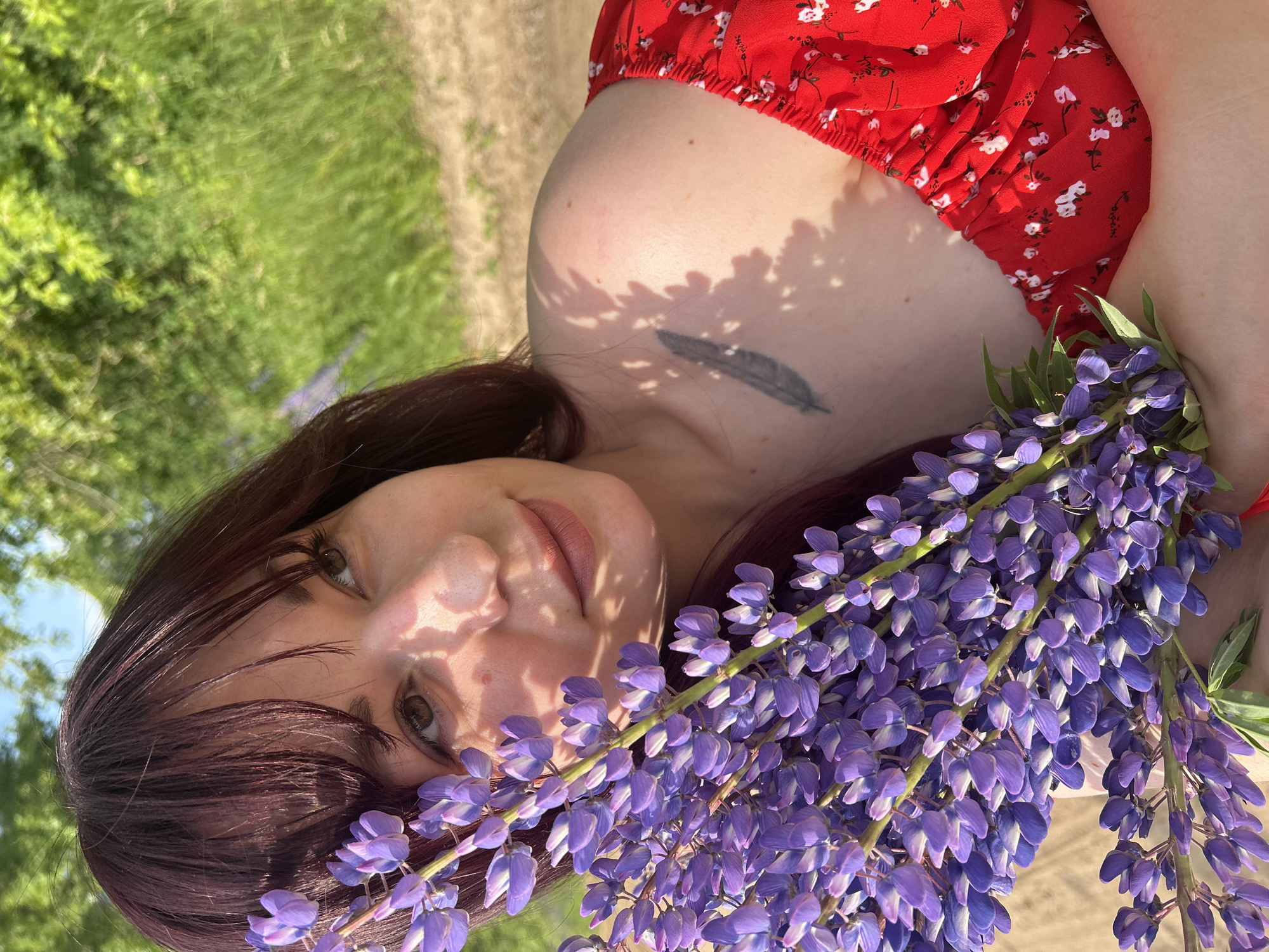 A young woman with dark hair and fair skin is holding a bouquet of purple lupine flowers. She is wearing a red dress with a white floral pattern. Her face is partially obscured by the flowers, but she appears to be looking away from the camera with a gentle smile. The background consists of blurred green foliage and a dirt path, suggesting an outdoor setting in a natural environment. There is a feather tattoo visible on her upper chest. The image captures a moment of natural beauty and tranquility, likely in a rural or park-like area. The specific location in the city of Гаревая, Russia is not discernible from the visual information provided.