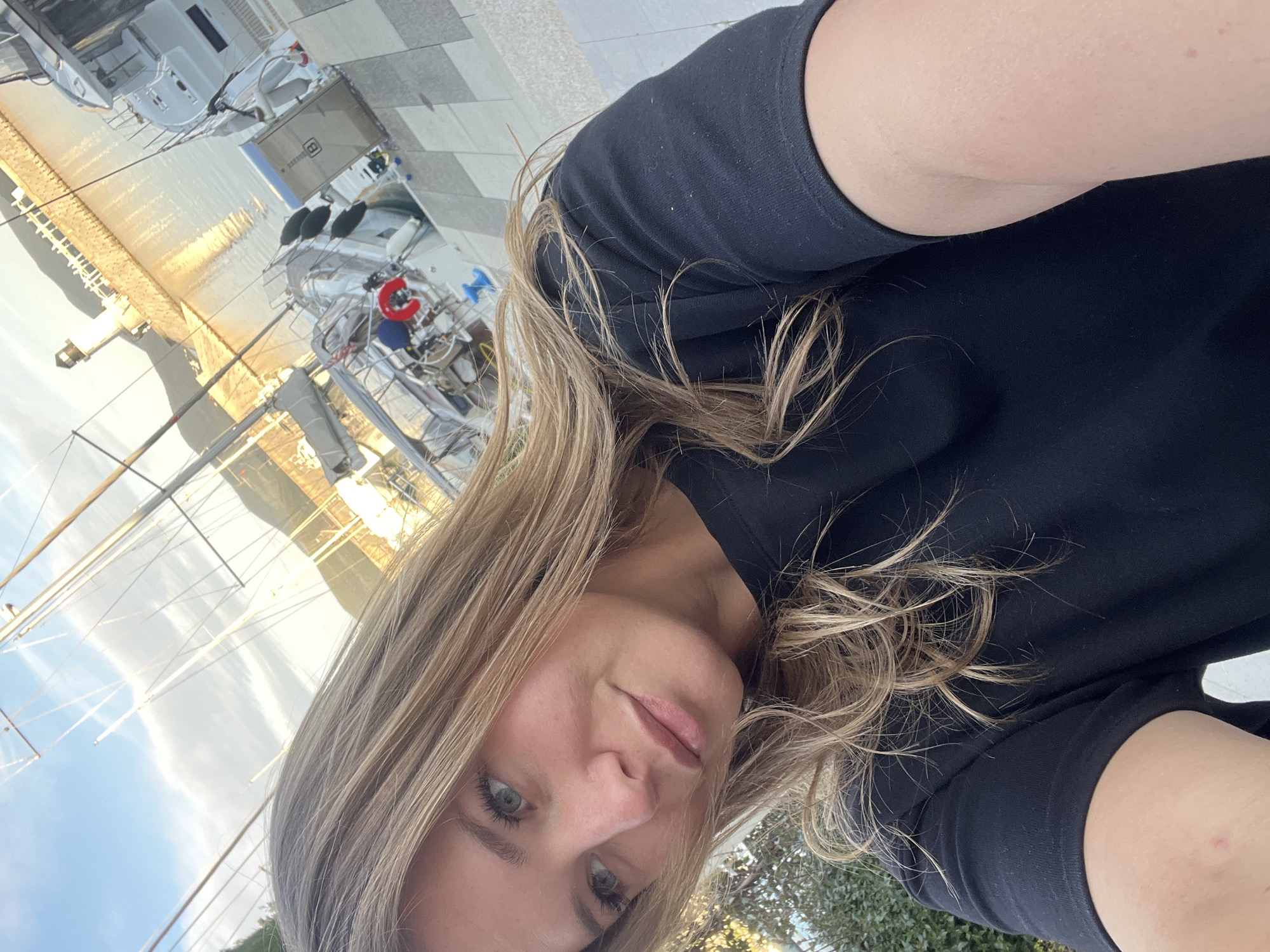 The image is a selfie-style portrait of a woman set against the backdrop of a sunny marina. The photograph is oriented at an angle, with the woman's face and upper body occupying the foreground. She has long, wavy blonde or light brown hair that cascades over her shoulders and back. She is wearing a plain black t-shirt or top. Her eyes are a striking blue-grey, looking directly into the camera, and her lips are closed with a subtle, serene expression.
In the background, numerous white yachts and sailboats are docked in a marina. The water reflects a warm, golden light, suggesting either an early morning or late afternoon sun. Masts, rigging, and various boat details such as railings, a red lifebuoy, and dark fenders are visible. On one of the boats, a grey rectangular container is noticeable, featuring a white letter or number, possibly 'B' or '8'. The marina area includes a paved walkway, and some green foliage is visible along the right edge of the frame. The sky is bright and clear, indicating fair weather. The setting is a tranquil coastal environment, consistent with a marina in Radovići, Montenegro.