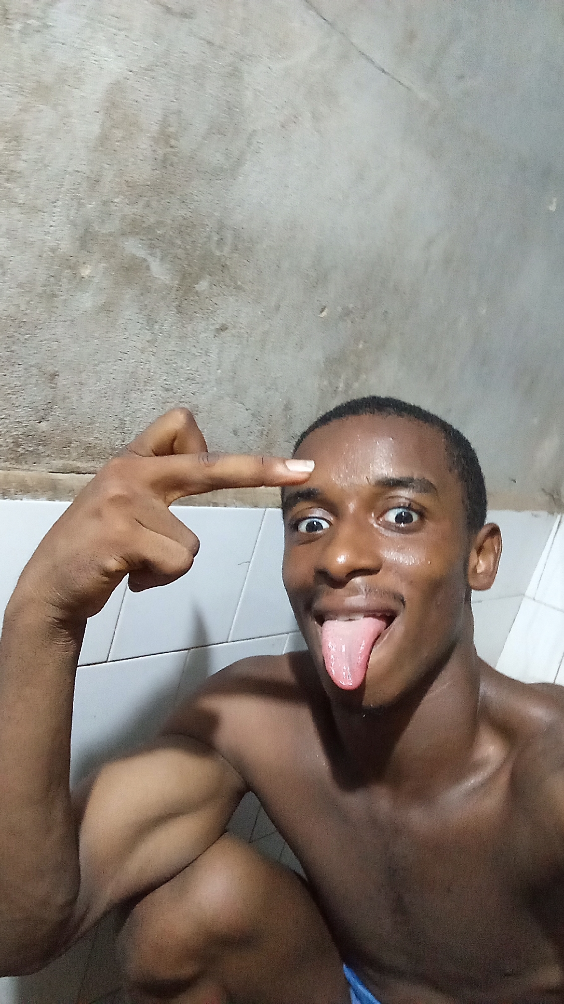 A young man with dark skin and short, dark hair is captured in a close-up, expressive pose, appearing to be in an indoor setting. He is shirtless, revealing a toned upper body with visible muscles, and his skin appears to be glistening, possibly from sweat or water.
His face is highly animated; he has wide, bright eyes and his tongue is fully extended out of his mouth in a playful or silly gesture. His left hand is raised to his head, with his index finger pointing directly at his forehead, while the other fingers are bent. This gesture, combined with his facial expression, conveys a humorous and goofy mood, possibly indicating a "mind blown" or "thinking" pose.
The background consists of two distinct sections. The upper portion is a rough, textured wall with a concrete-like appearance, showing variations in grey and brown tones. Below this, a section of white rectangular tiles with dark grout lines is visible, suggesting a utilitarian space like a bathroom or a utility room. The lighting appears somewhat dim but sufficient to highlight the subject. There is no visible text in the image.