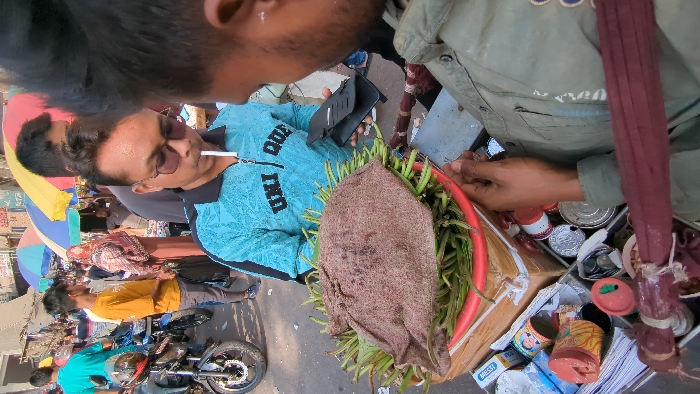 The image displays a bustling street market scene in Dhaka, Bangladesh, under bright daylight.
In the immediate foreground, the right arm and shoulder of an individual wearing an olive-green long-sleeved shirt are partially visible on the right. This individual extends their right hand towards a red plastic basket. This basket, serving as a vendor's display, is filled with green elongated vegetables, possibly string beans, and partially covered by a coarse, textured brown fabric. The basket rests on a wooden crate, beneath which a compartment of a vendor's cart contains various small consumer goods, including metal cans with labels, a red plastic container, and stacked papers.
Opposite this, a man with short dark hair and dark sunglasses is centrally featured. He wears a turquoise blue long-sleeved shirt with the text "UNI DUE" visible on the chest. A white cigarette is held in his mouth, and his left hand clutches a black mobile phone case. He appears to be engaging with the vendor's goods.
The background reveals a crowded urban environment. Numerous pedestrians are visible, including a man in a bright yellow t-shirt standing near parked motorcycles on the left, and a woman in a patterned head covering and brown dress further back. Several motorcycles are parked along the street. Colorful striped market umbrellas (red, blue, yellow) provide shade for various stalls. The ground is paved, and indistinct buildings line the street in the distance.